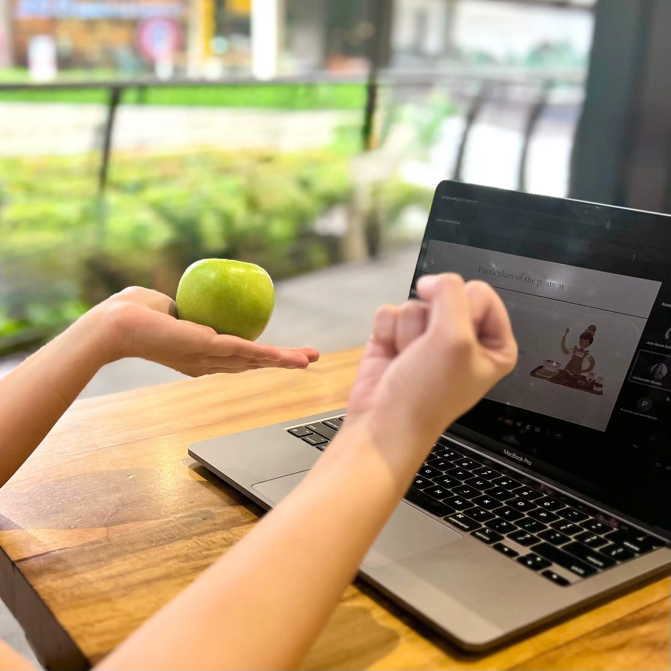 A dietitian holding a green apple next to a laptop on a wooden table, with a blurred garden view in the background.