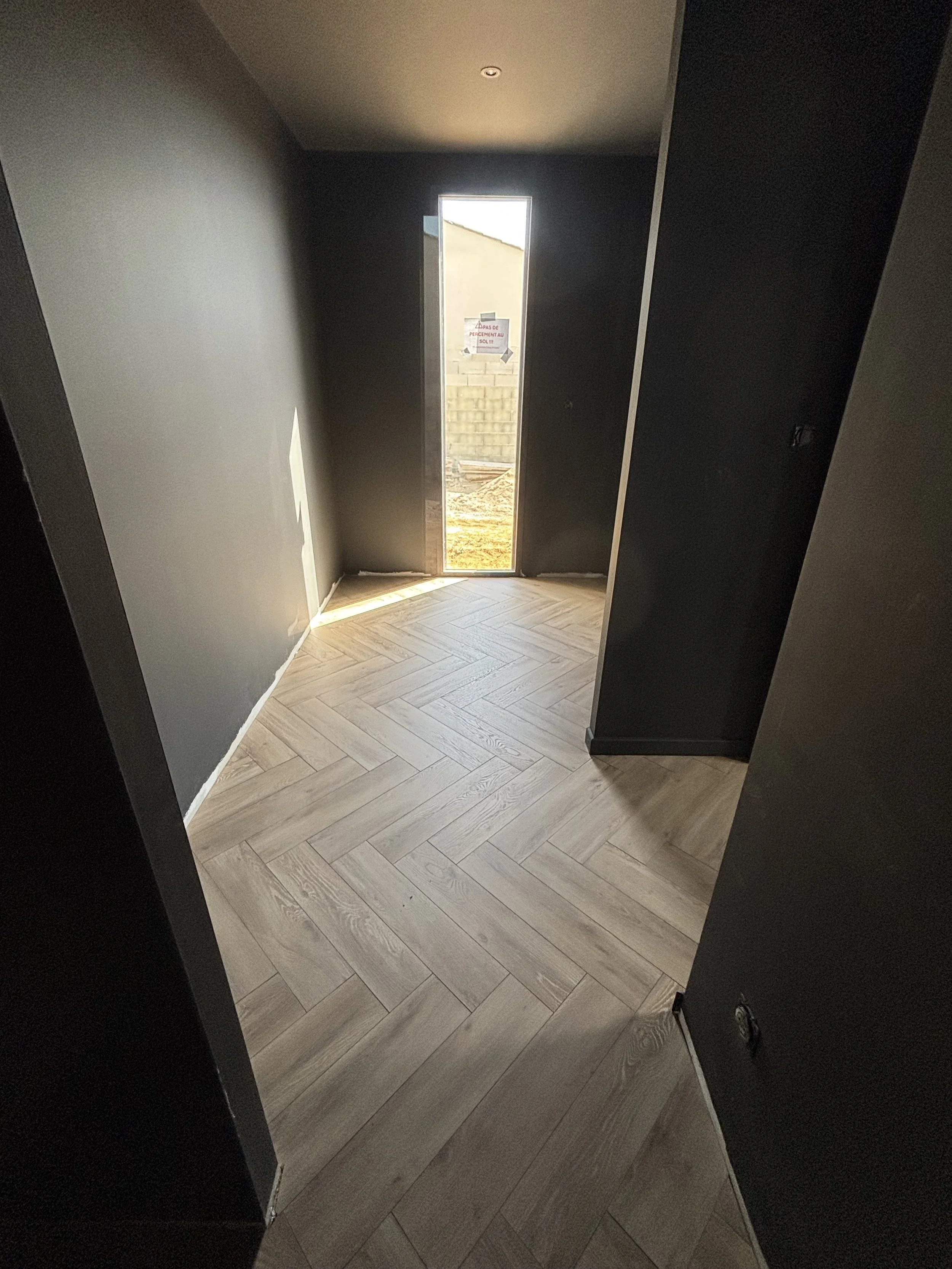 Intérieur d'une pièce avec murs noirs, sol en bois clair en motif parquet et une porte en position verticale laissant entrer la lumière du soleil, vue vers l'extérieur avec un mur en briques au fond.