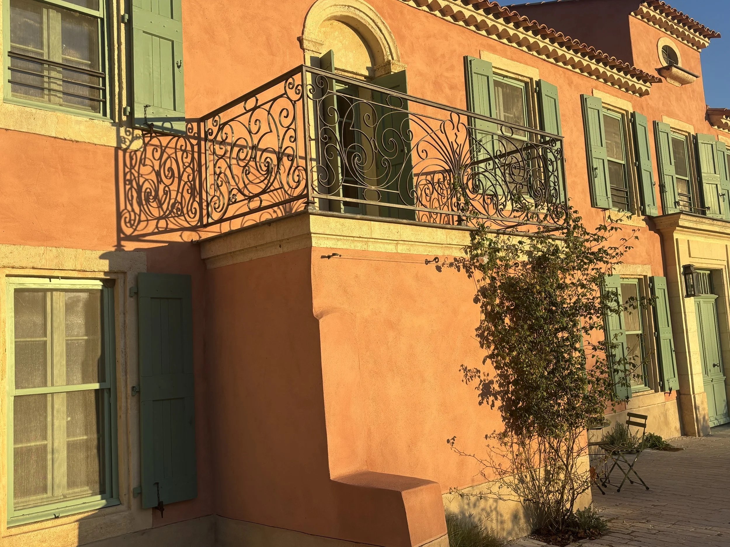 Une façade de maison colorée avec des fenêtres à volets verts, un balcon avec une rampe en fer forgé, un arbre devant la maison, et une chaise pliante noire.