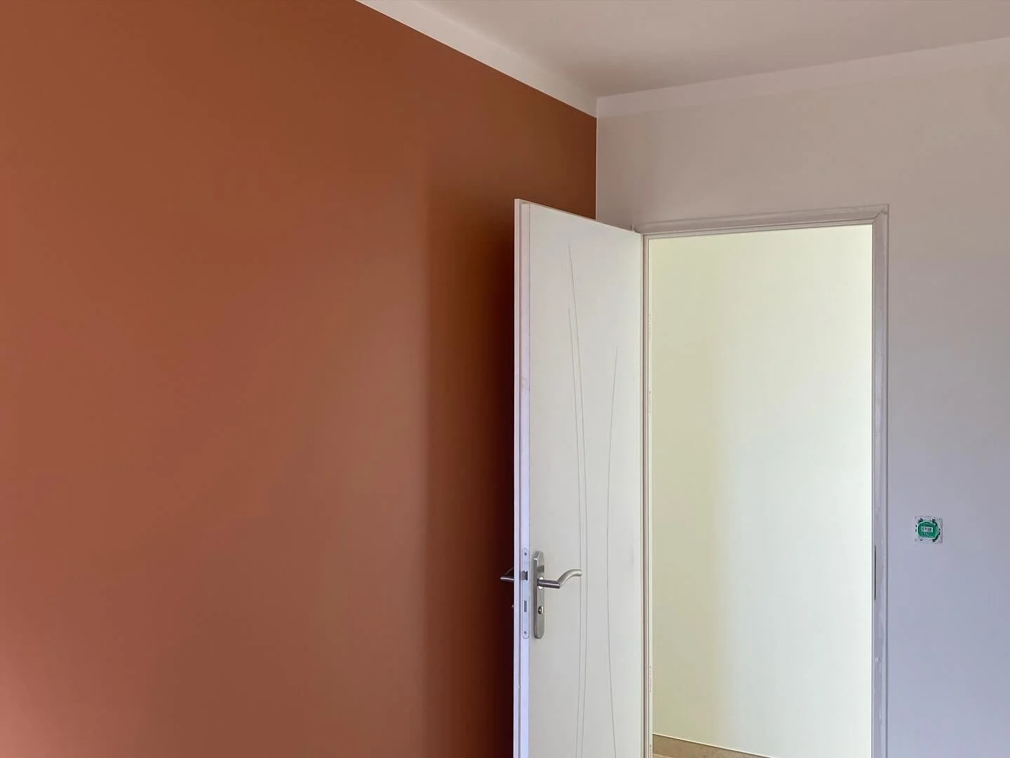 Une porte blanche partiellement ouverte dans une pièce avec un mur beige et un autre blanc, et une prise électrique verte à côté.