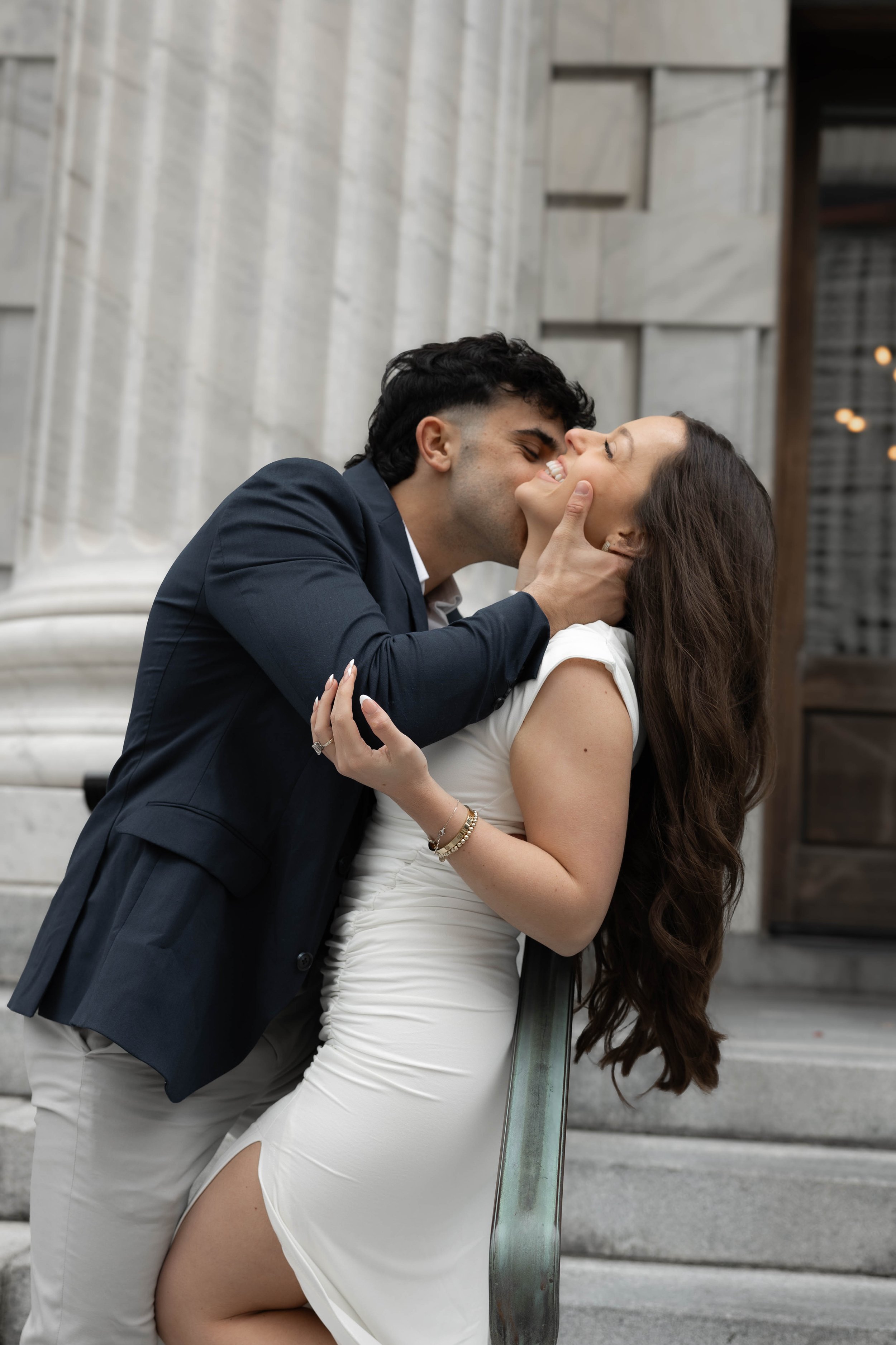 A couple sharing a romantic kiss on a staircase in front of a building with columns. The man is wearing a dark suit, and the woman is wearing a white dress.