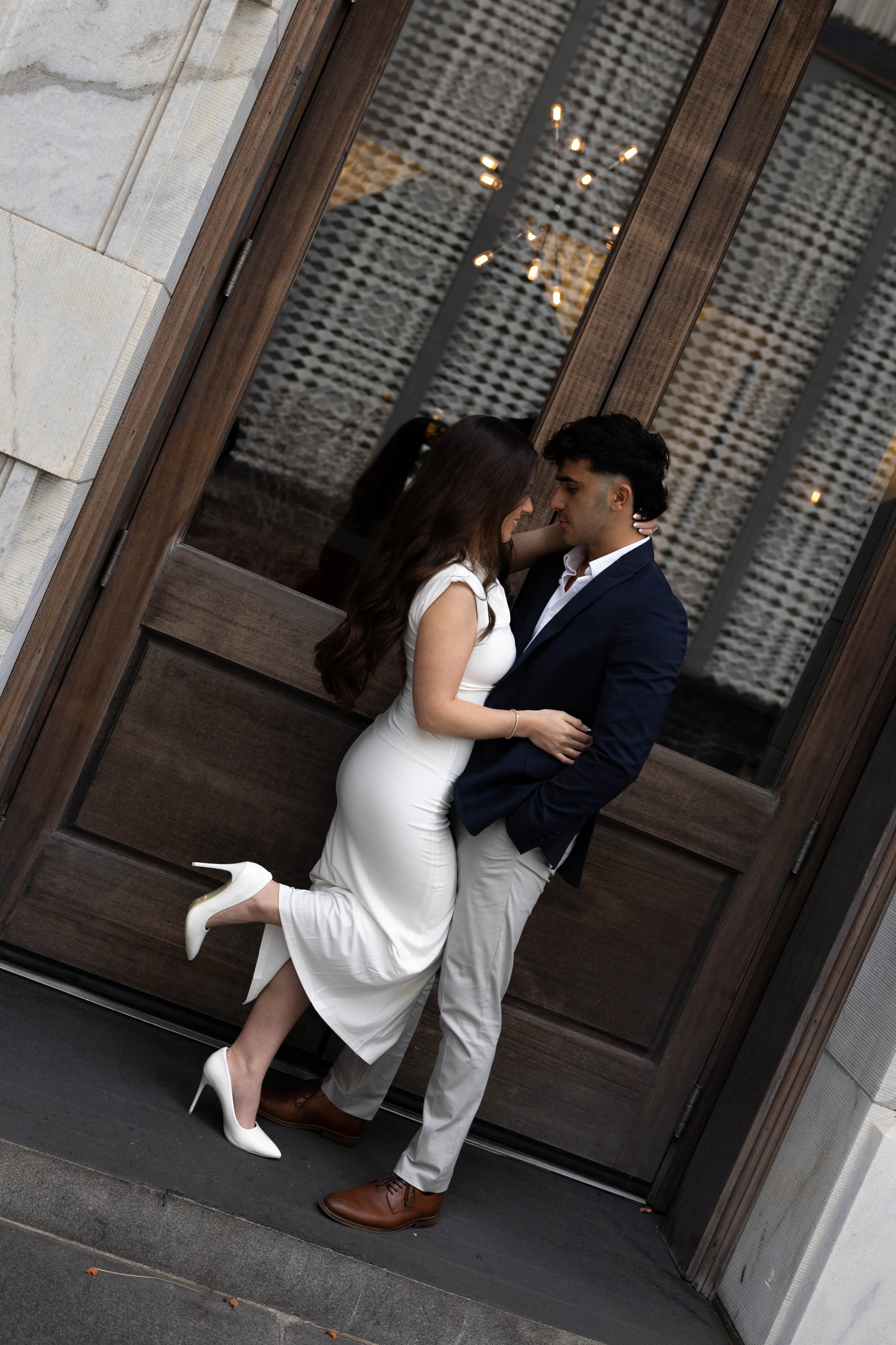 A couple embracing in front of a wooden door. The woman is wearing a white dress and white high heels, and the man is dressed in a navy blazer, white shirt, and beige pants.