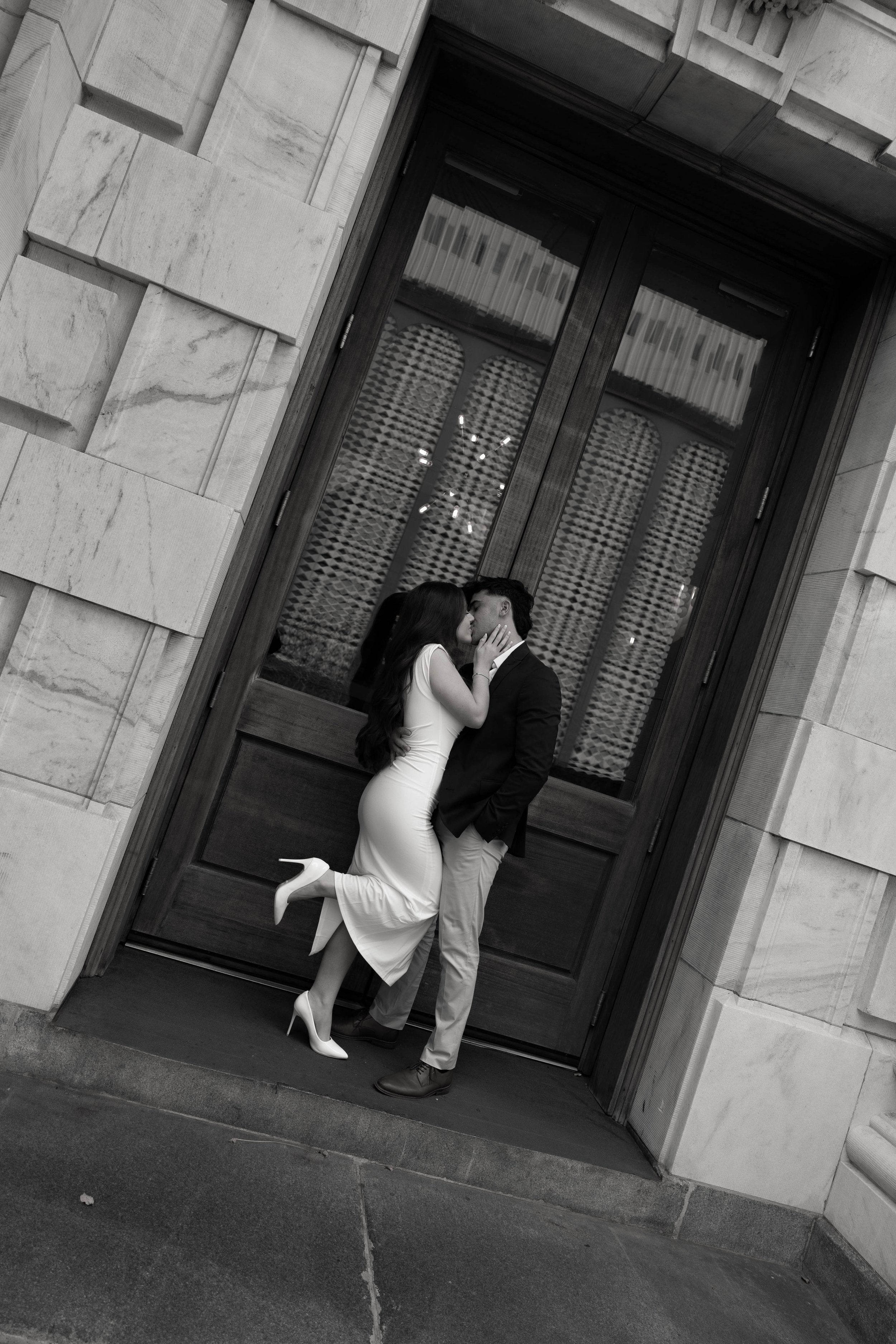 A couple kisses in front of a large wooden door, with a tall woman in a white dress and heels and a man in a dark jacket and light pants.