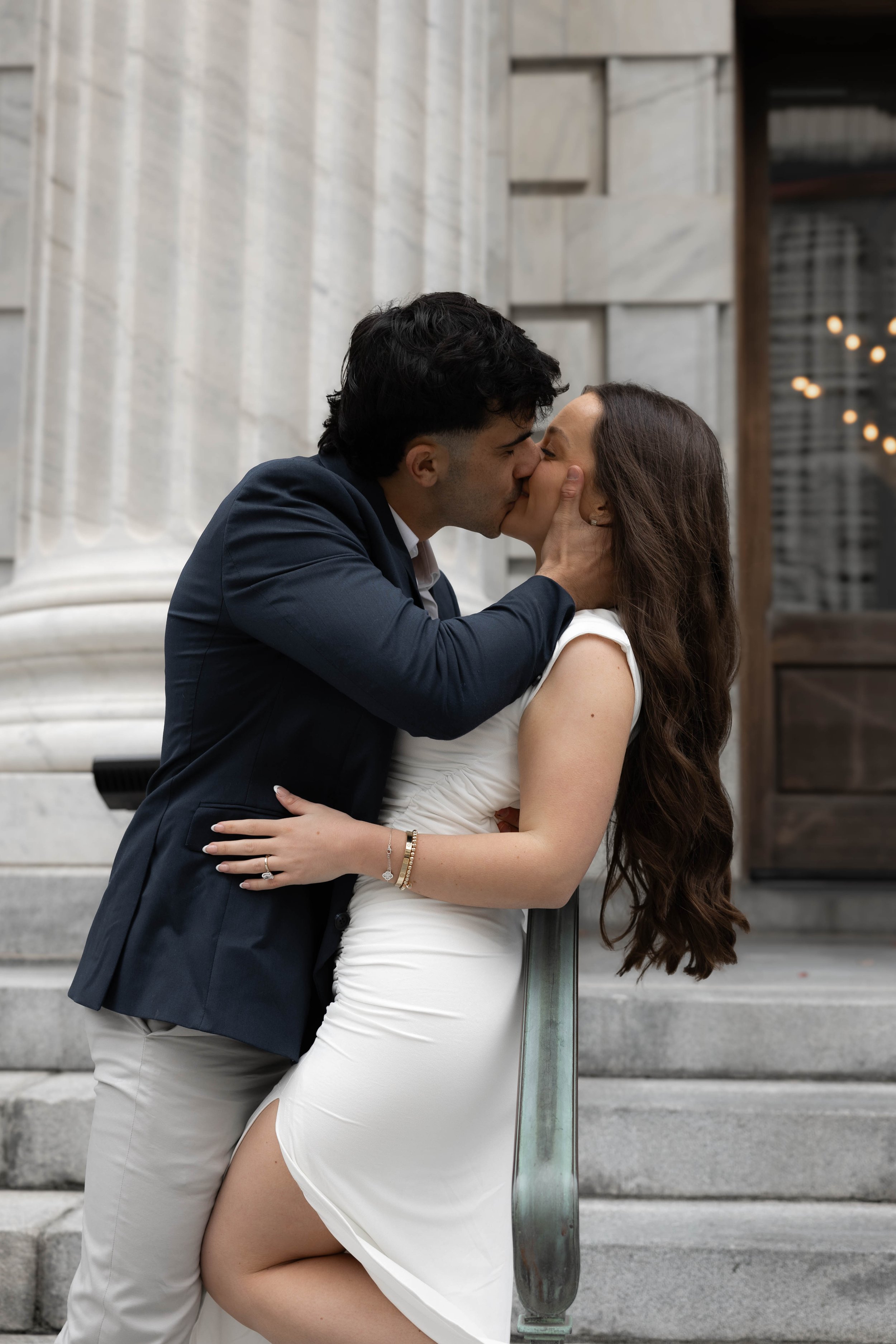 Couple kissing on stone steps, woman in white dress, man in dark jacket, architectural background.
