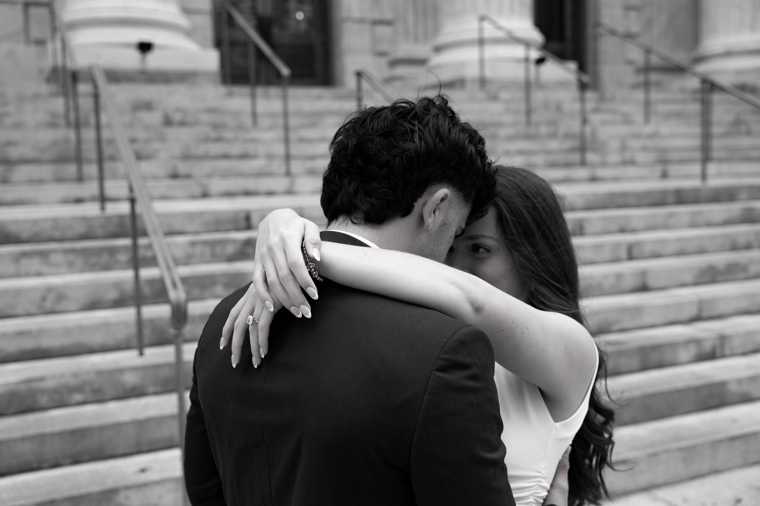 A couple embracing on steps, with the woman looking toward the camera and showing an engagement ring.