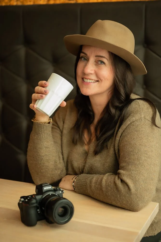 Frau mit Hut hält Tasse, sitzt an einem Tisch mit einer Kamera darauf.