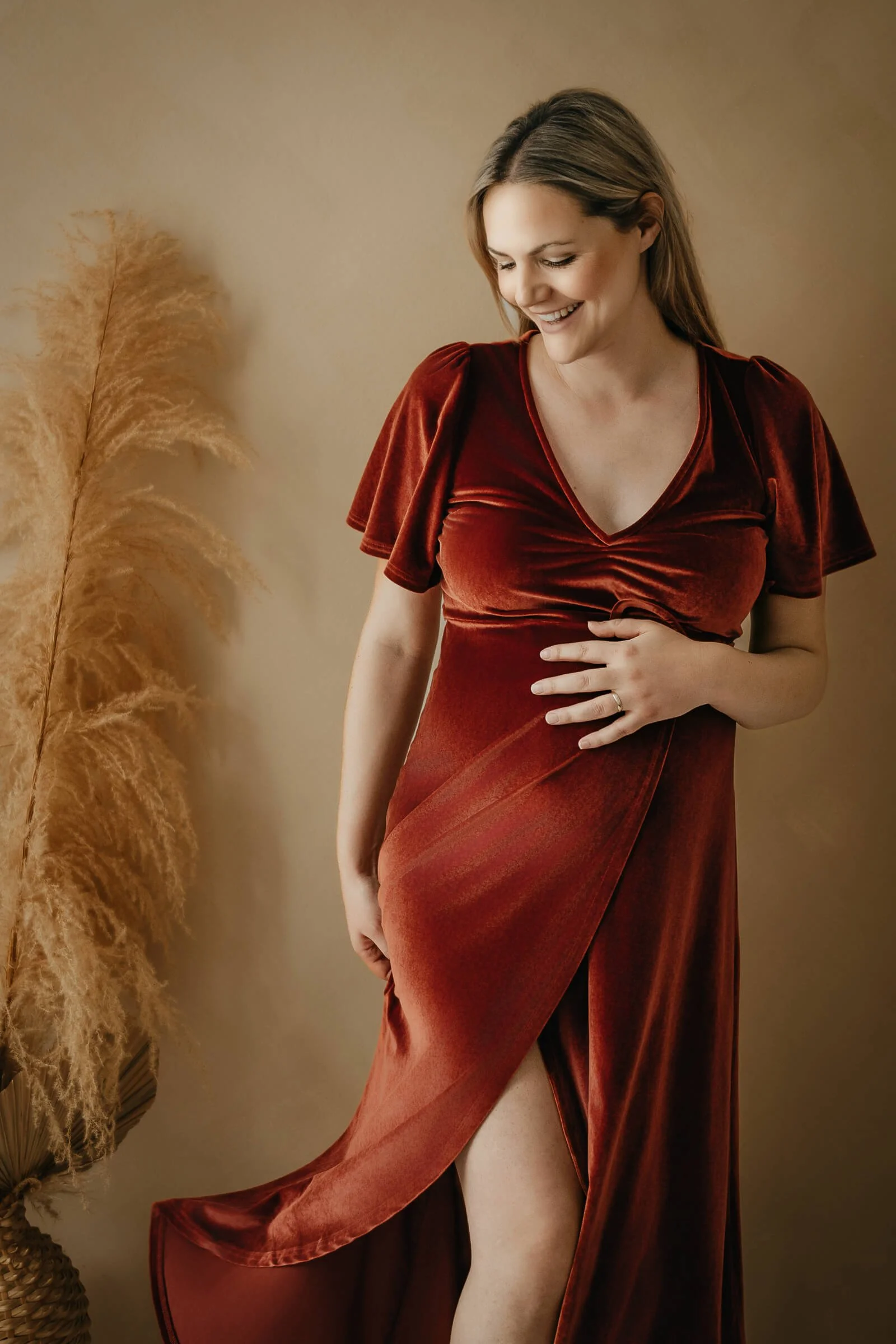 Eine lächelnde Frau in einem roten Samtkleid steht vor einer weißen Wand, umgeben von Dekorationen aus getrockneten Pflanzen.