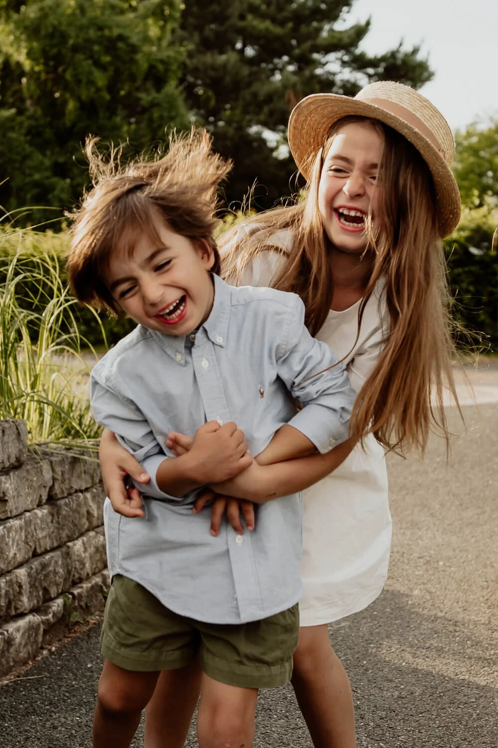 Zwei lachende Kinder spielen draußen, ein Junge in einem blauen Hemd und ein Mädchen mit Strohhut. Sie umarmen sich und wirken fröhlich.