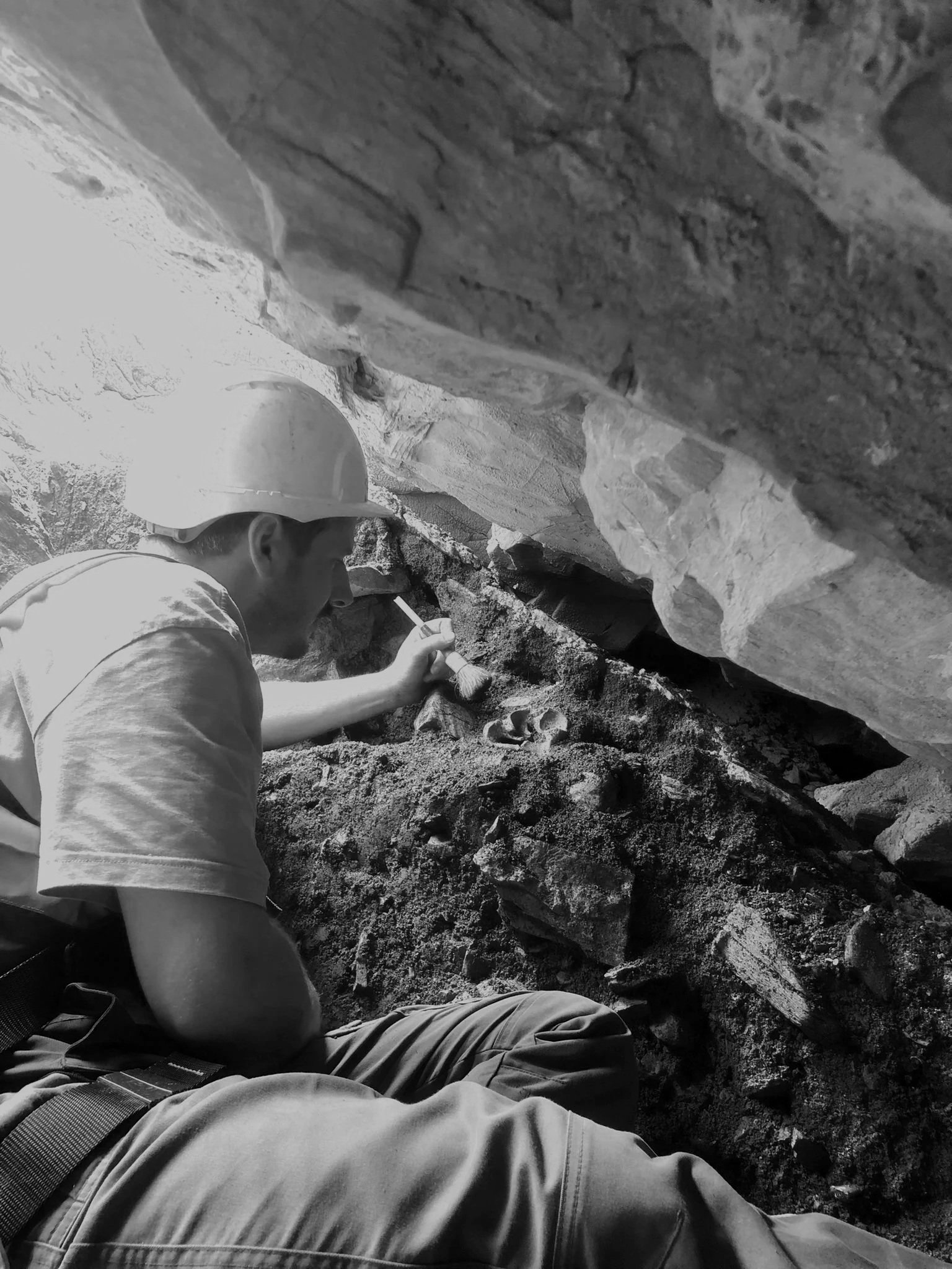 A person wearing a safety helmet and casual clothing, working on the ground in a rocky, outdoor environment, possibly excavating or inspecting soil or rocks.