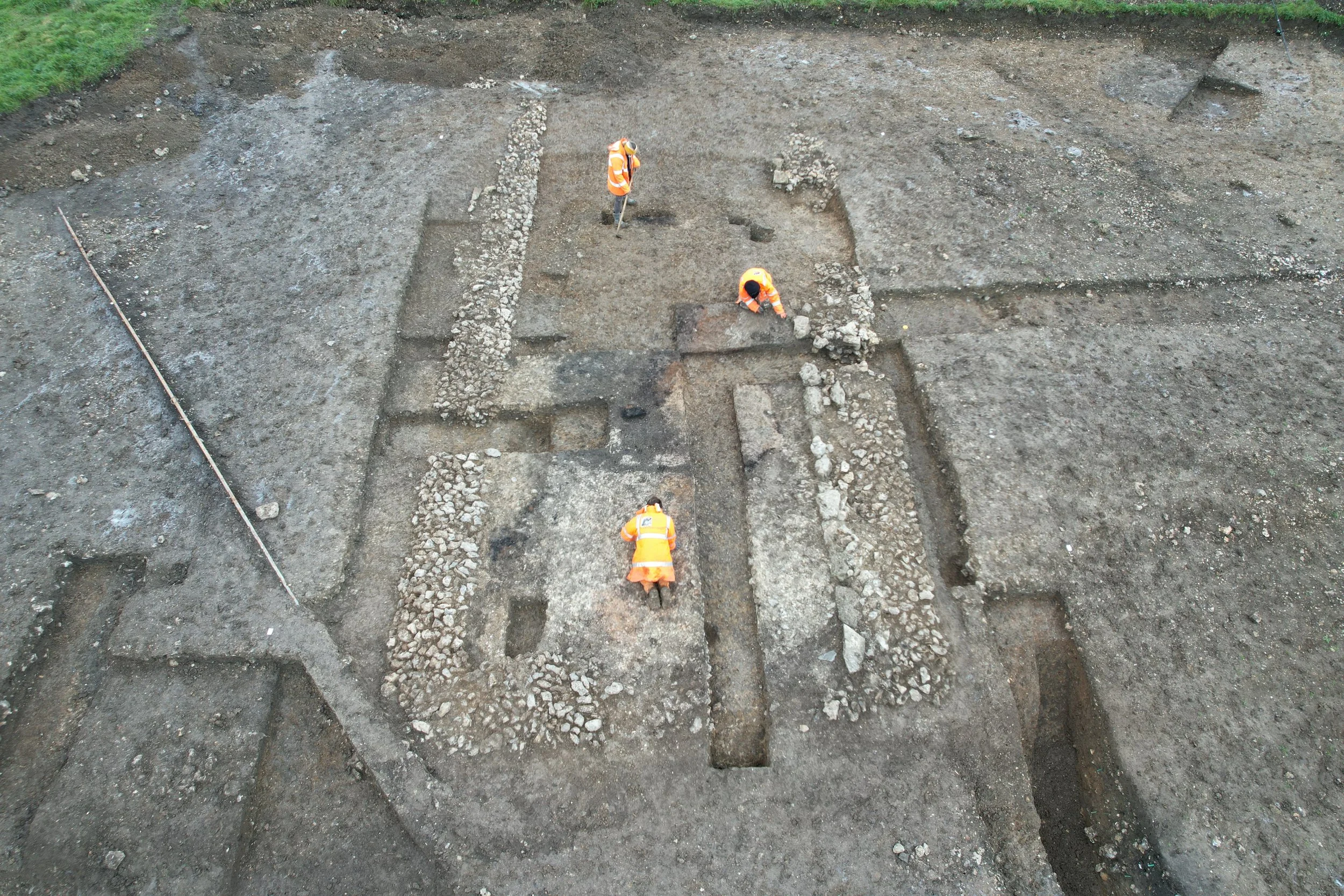 Three archaeologists in orange safety vests excavating an ancient site with stone structures.