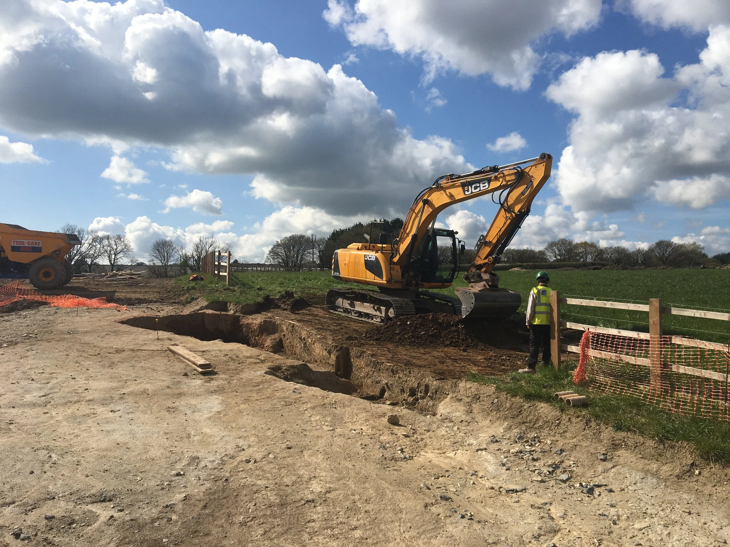 Construction worker in orange and black jacket using a shovel on a dirt site next to a teal excavator at a construction site.