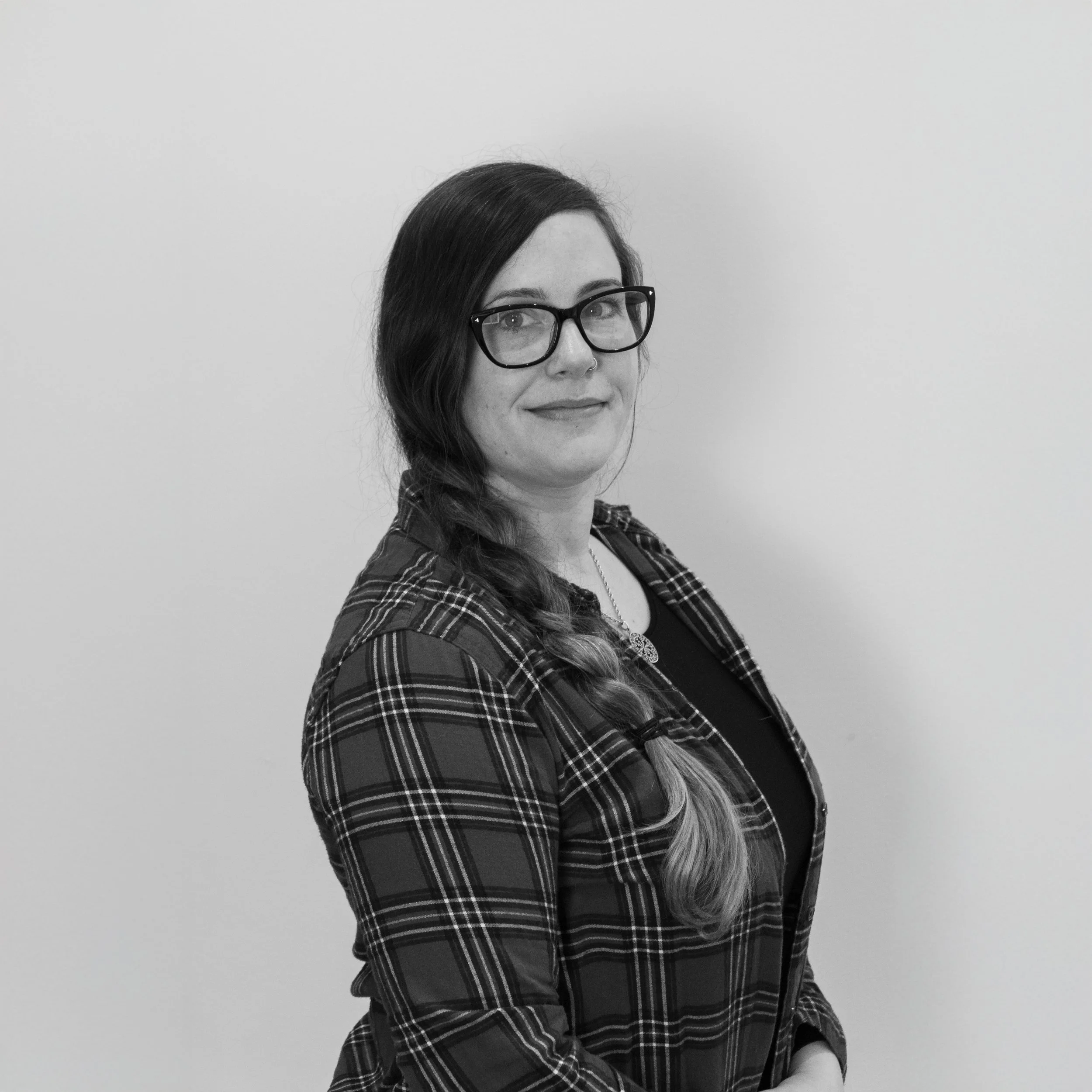A woman with long hair in a braid, wearing glasses, a necklace, and a plaid shirt, standing against a plain background.