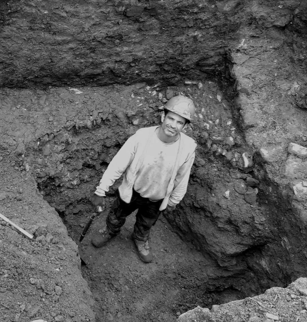 A man standing in a large excavation site, wearing a hard hat and work clothes, holding a tool, surrounded by dirt and soil.