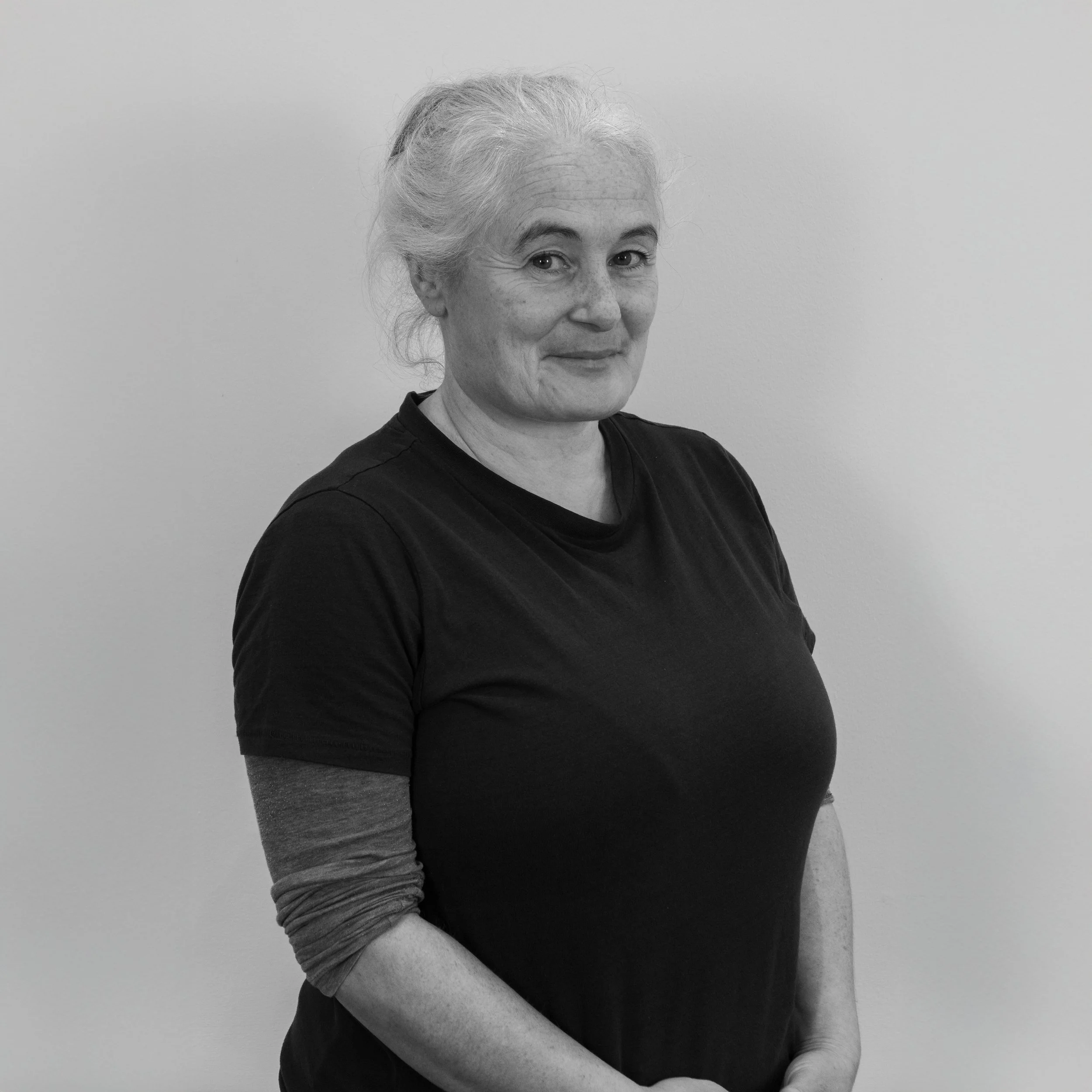 Black and white photo of a smiling woman with gray hair, wearing a black t-shirt and a long-sleeved shirt underneath, standing against a plain wall.