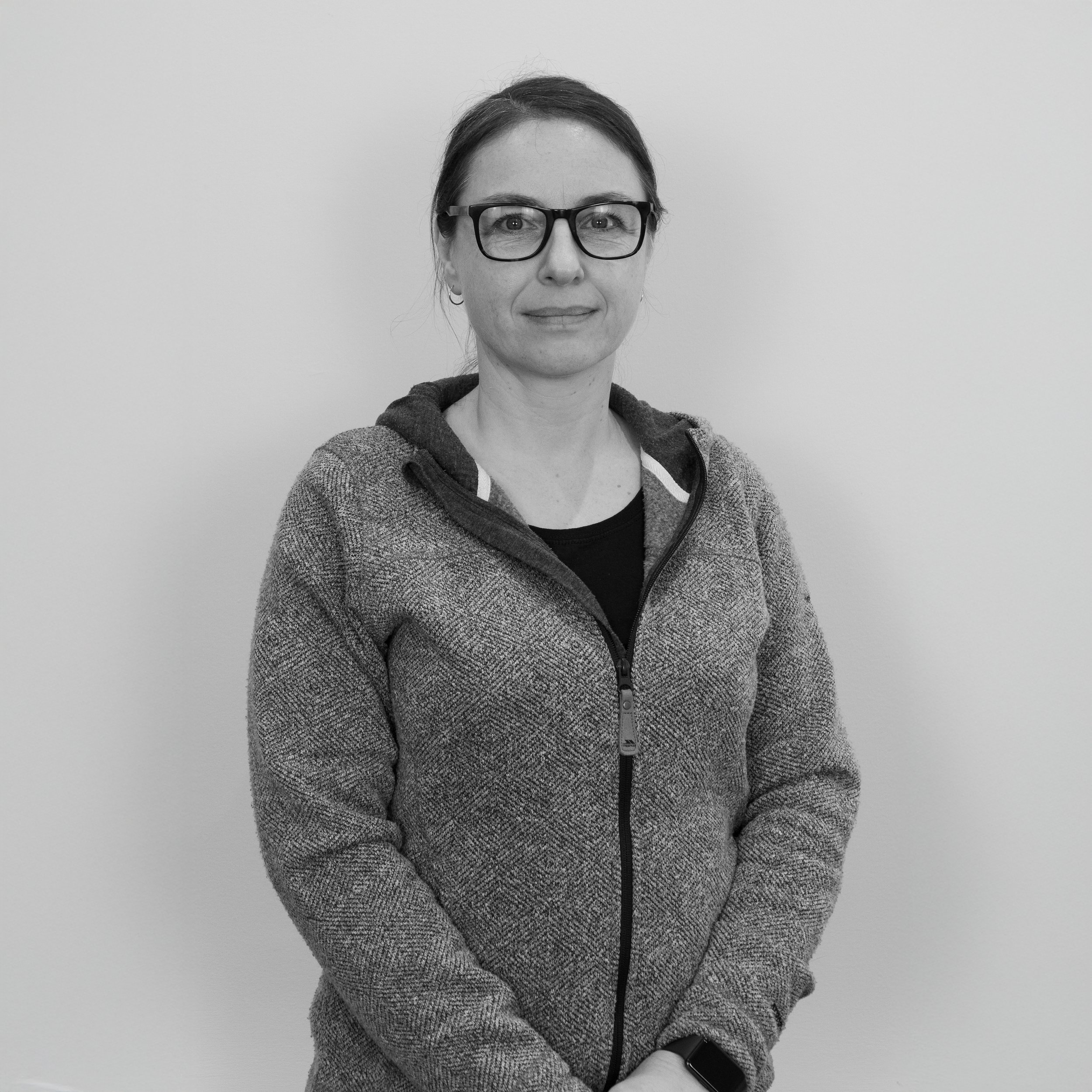 Black-and-white photo of a woman wearing glasses, a hoodie, and a smartwatch, standing against a plain background.