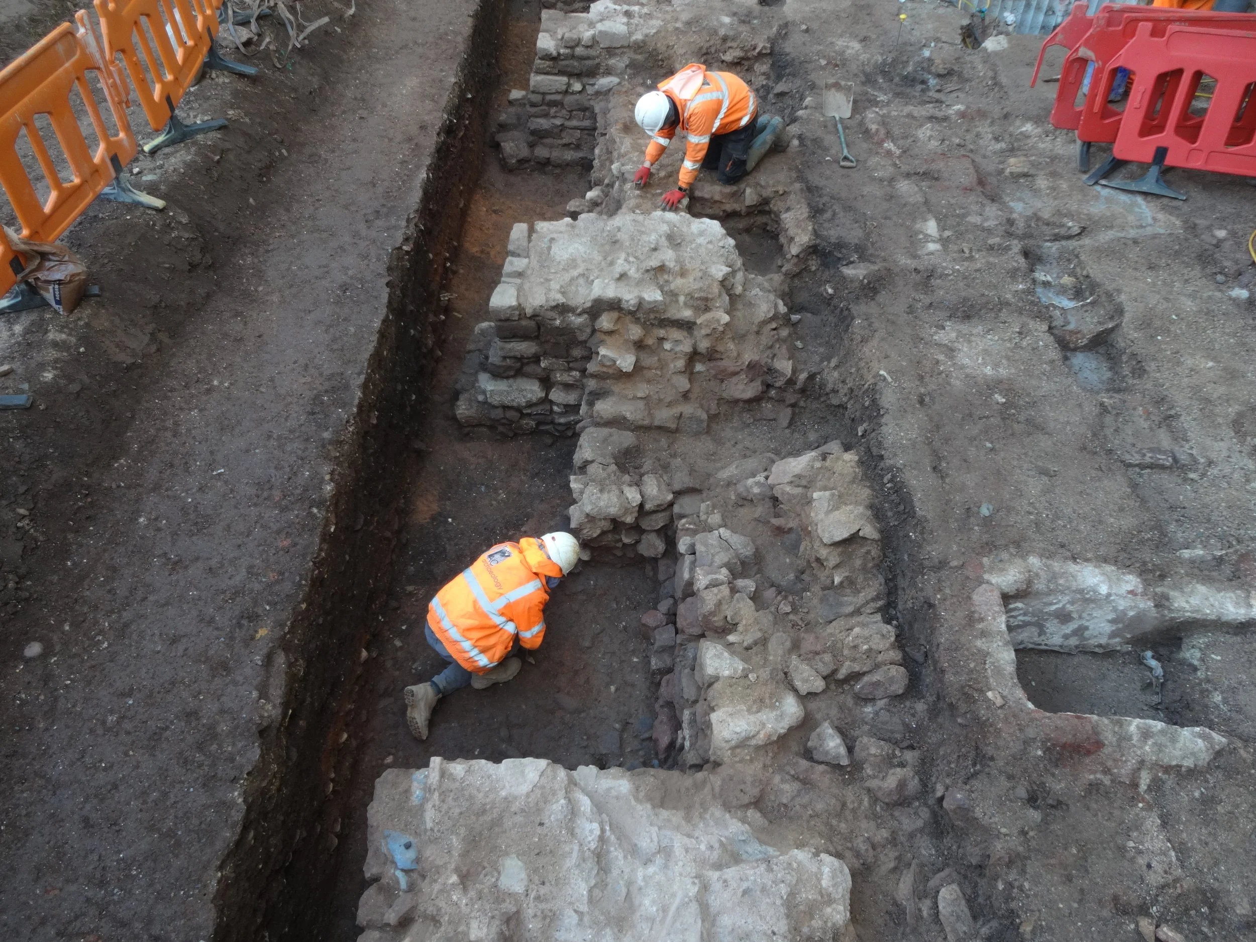 Two construction workers in orange safety jackets burying an artifact or historical stones at an archaeological excavation site, surrounded by disturbed dirt and temporary orange barriers.