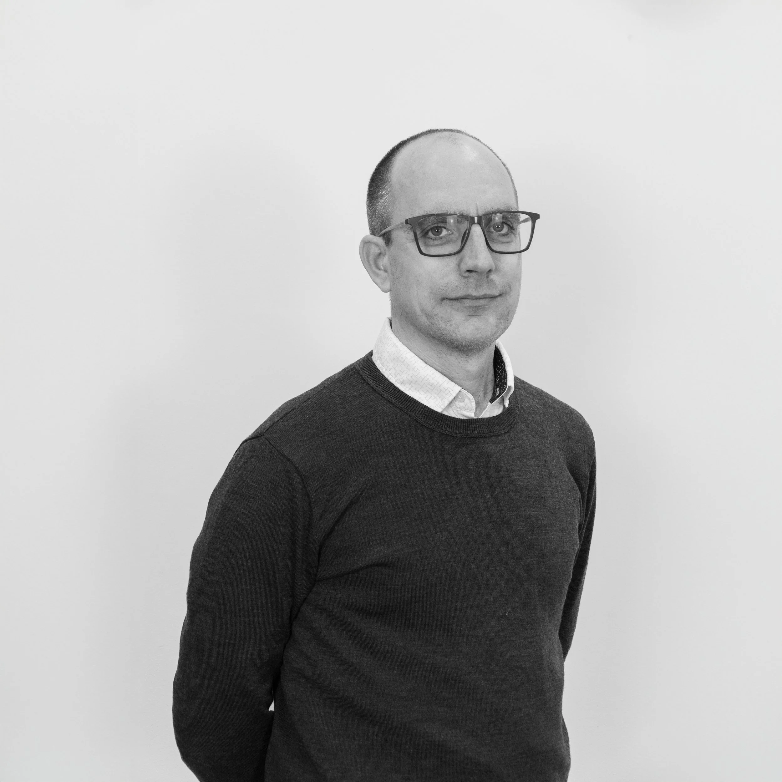 A black-and-white portrait of a man with glasses, a receding hairline, wearing a dark sweater over a collared shirt, standing against a plain white wall.