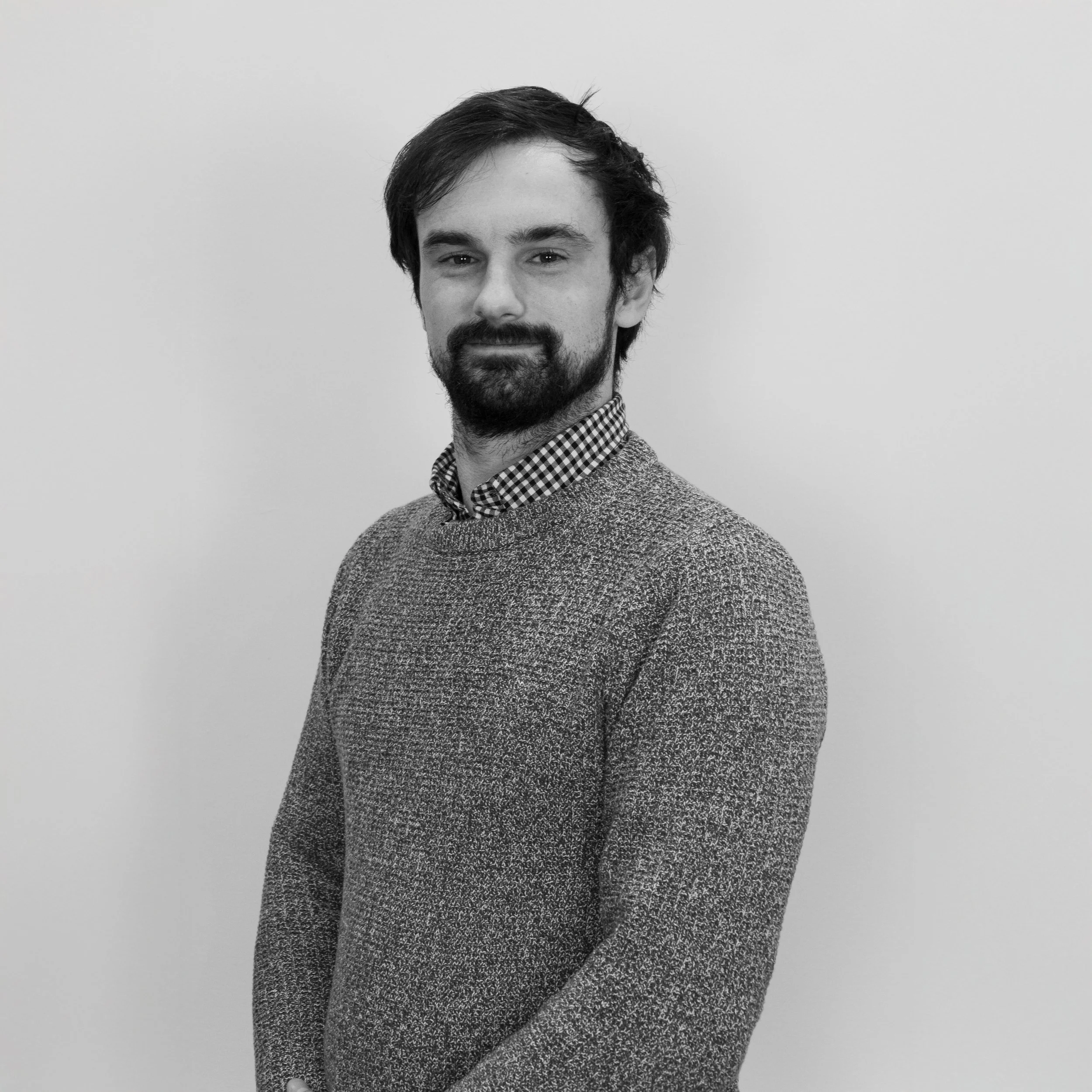 Black-and-white portrait of a man with dark hair, beard, wearing a patterned sweater over a checkered shirt, standing against a plain background.