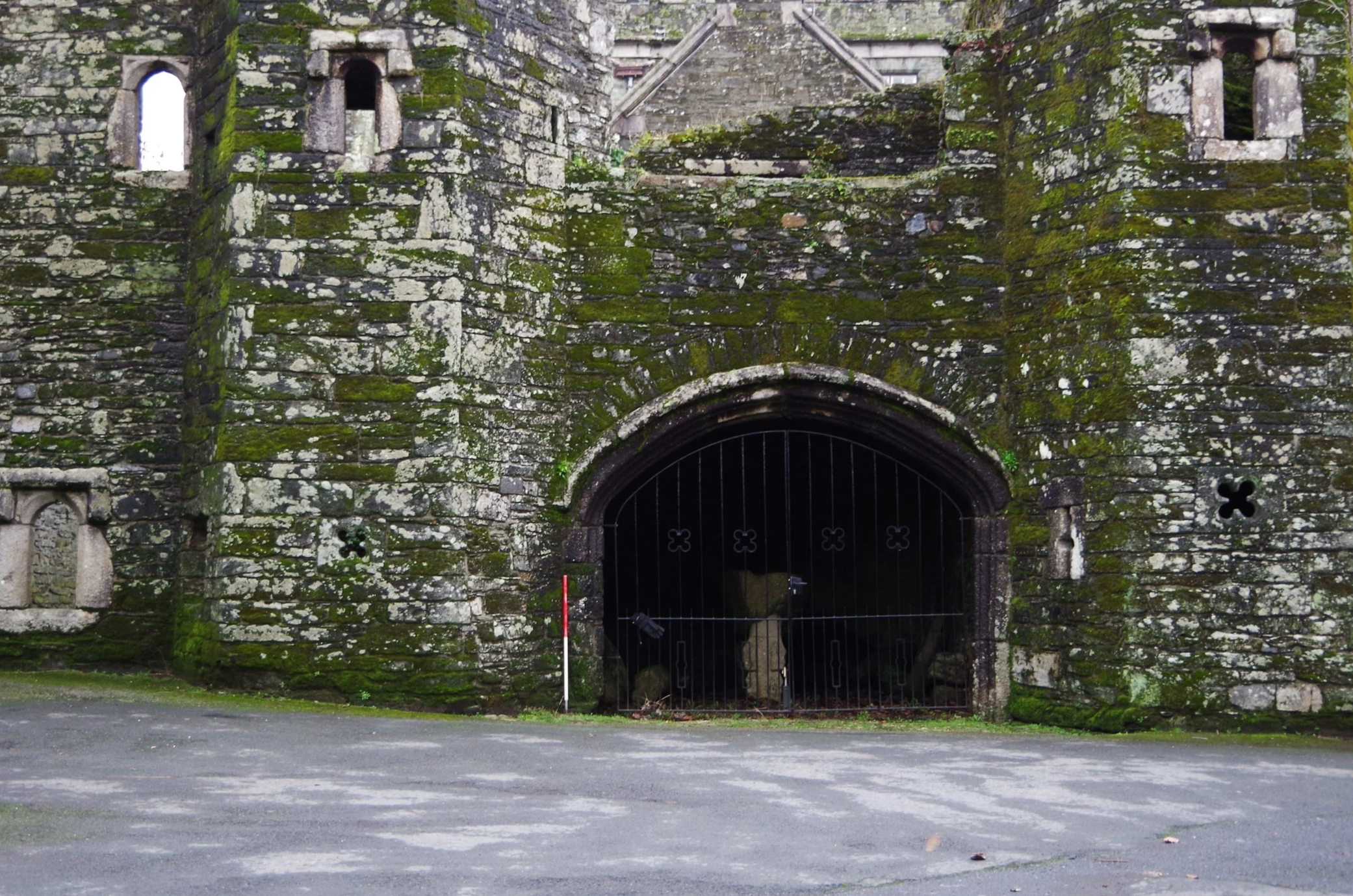 A large stone house with multiple chimneys and a bay window, surrounded by a green lawn and trees in the background, under a partly cloudy sky.