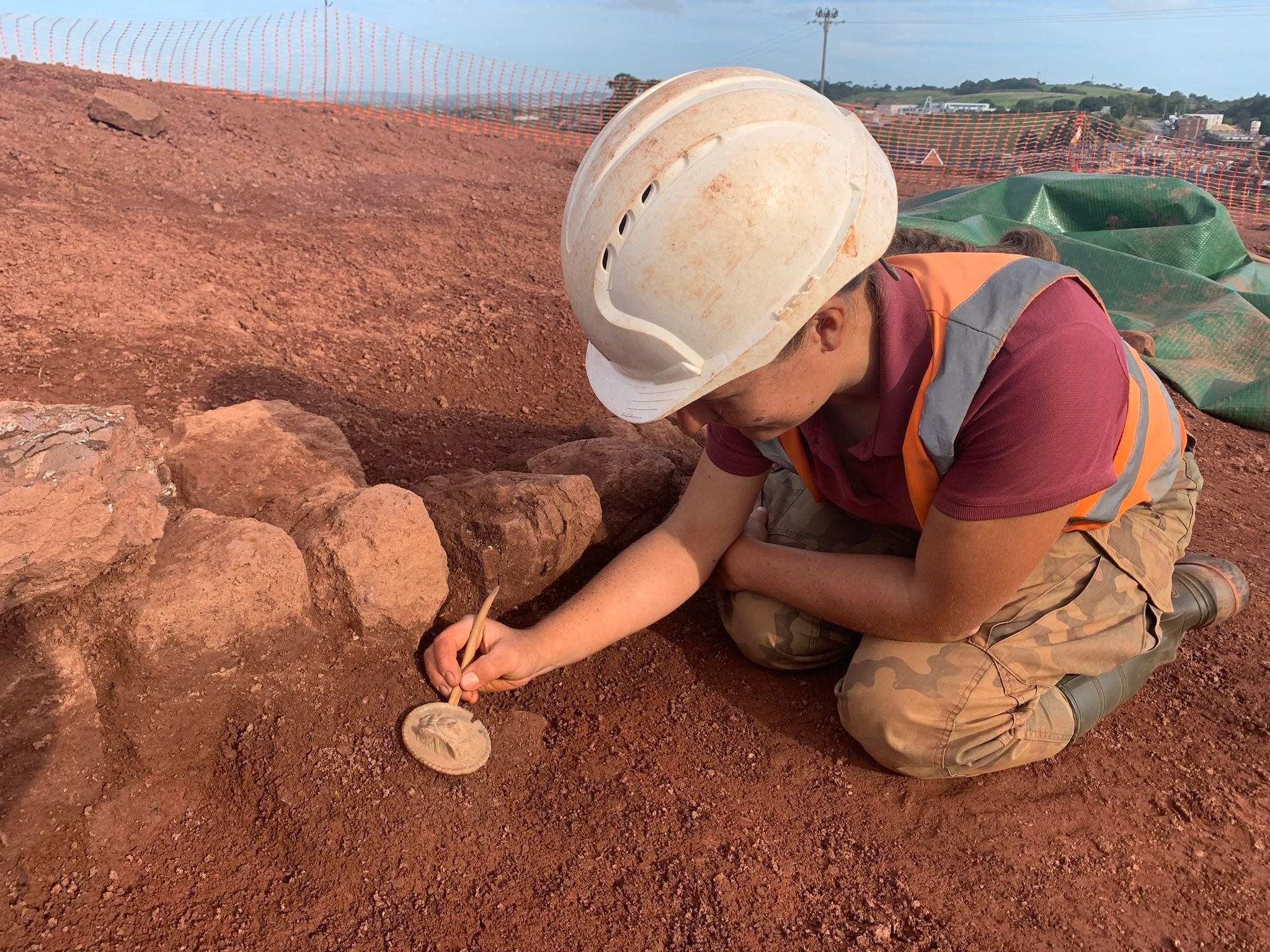 Four Archaeologists wearing safety helmets and orange vests working on a large, circular archaeological site with six rectangular excavations.