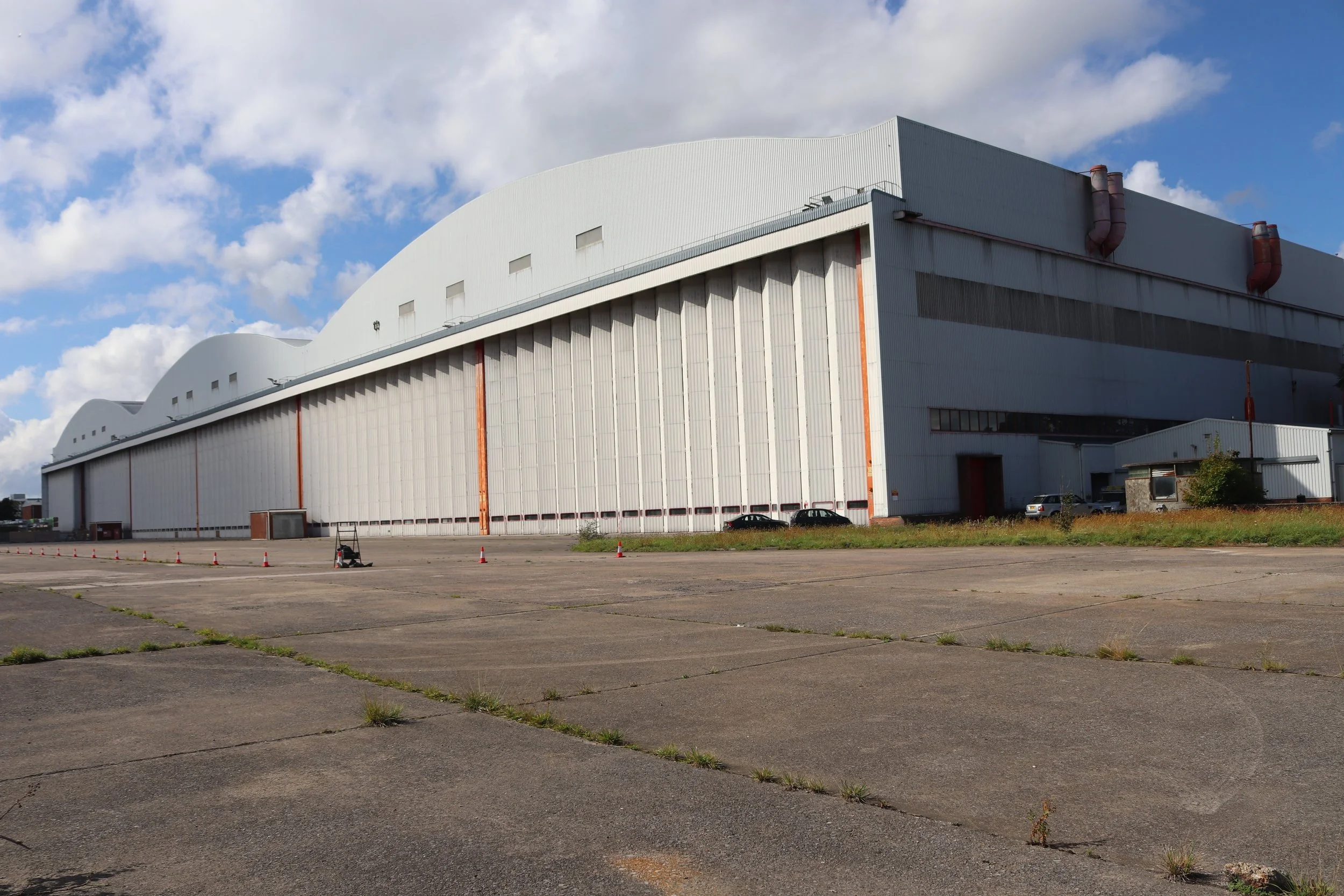 A large industrial building with a white, curved roof and tall, white walls, located in an open, paved area with some grass and parked cars.