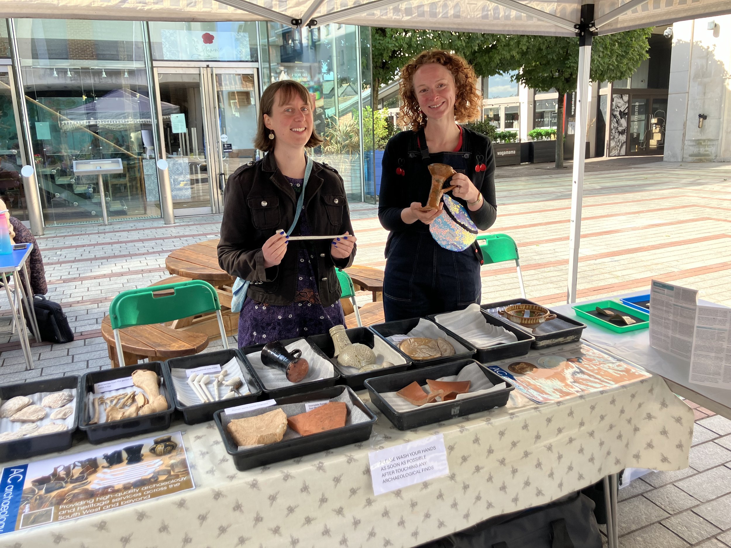 Two women standing behind a table displaying various archaeological artifacts, with one holding a stone tool and the other holding a ceramic piece, under a white canopy outdoors in a plaza.