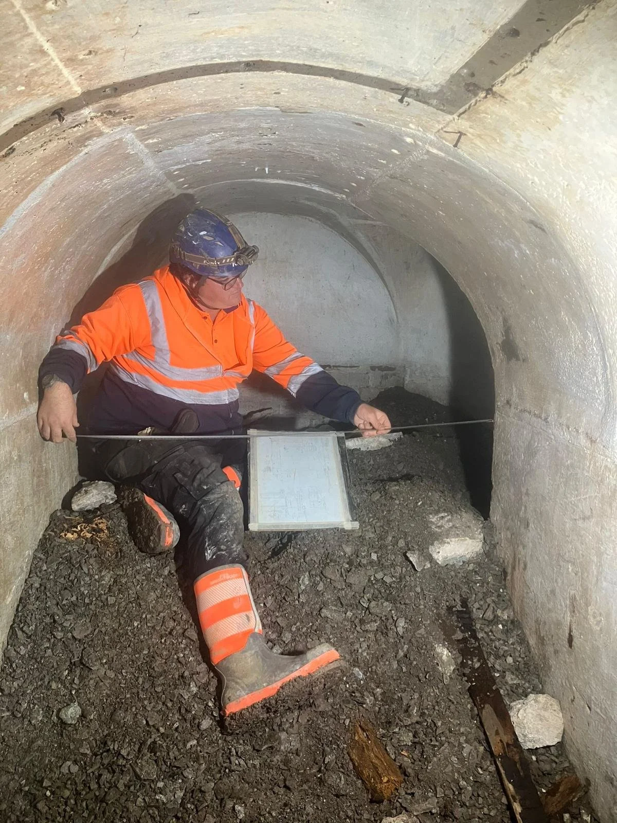 Worker in safety gear inspecting the floor inside a tunnel with a flashlight and a measurement device