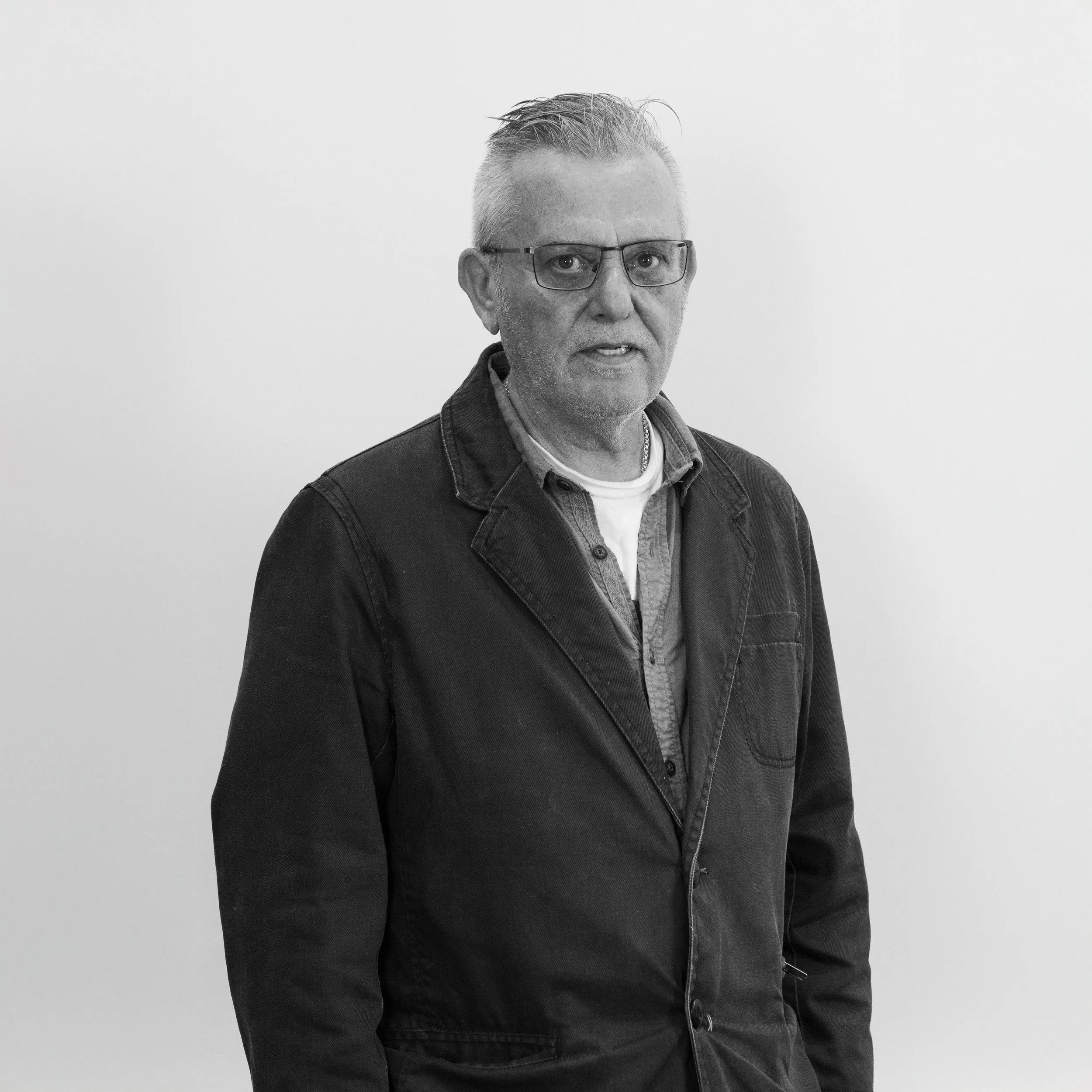 Black and white photo of a middle-aged man with glasses, short and styled hair, wearing a dark jacket over layered shirts, standing against a plain light-colored wall.