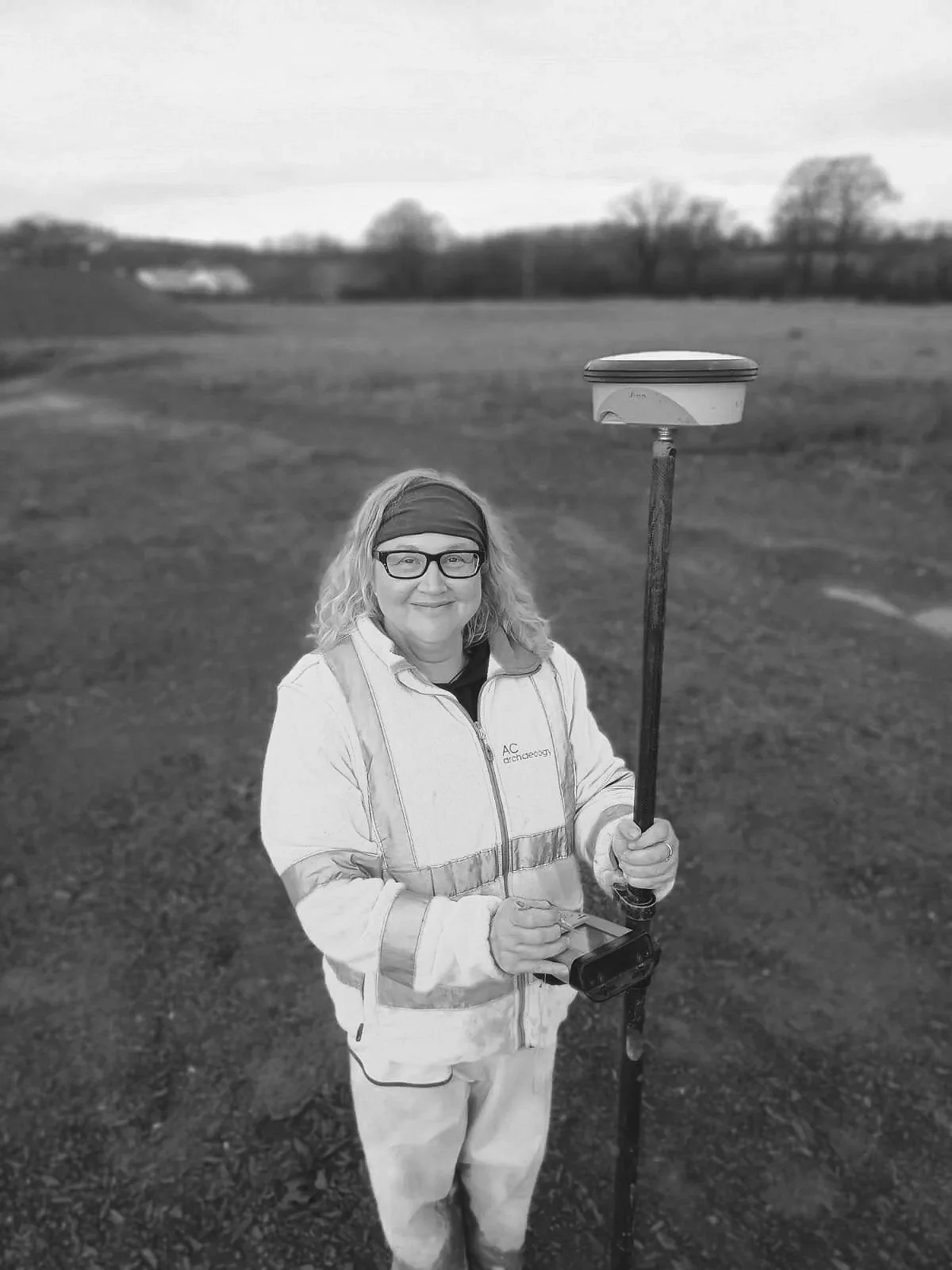 A woman standing outdoors in a field, holding a surveying instrument on a tripod with one hand and a device in the other hand.
