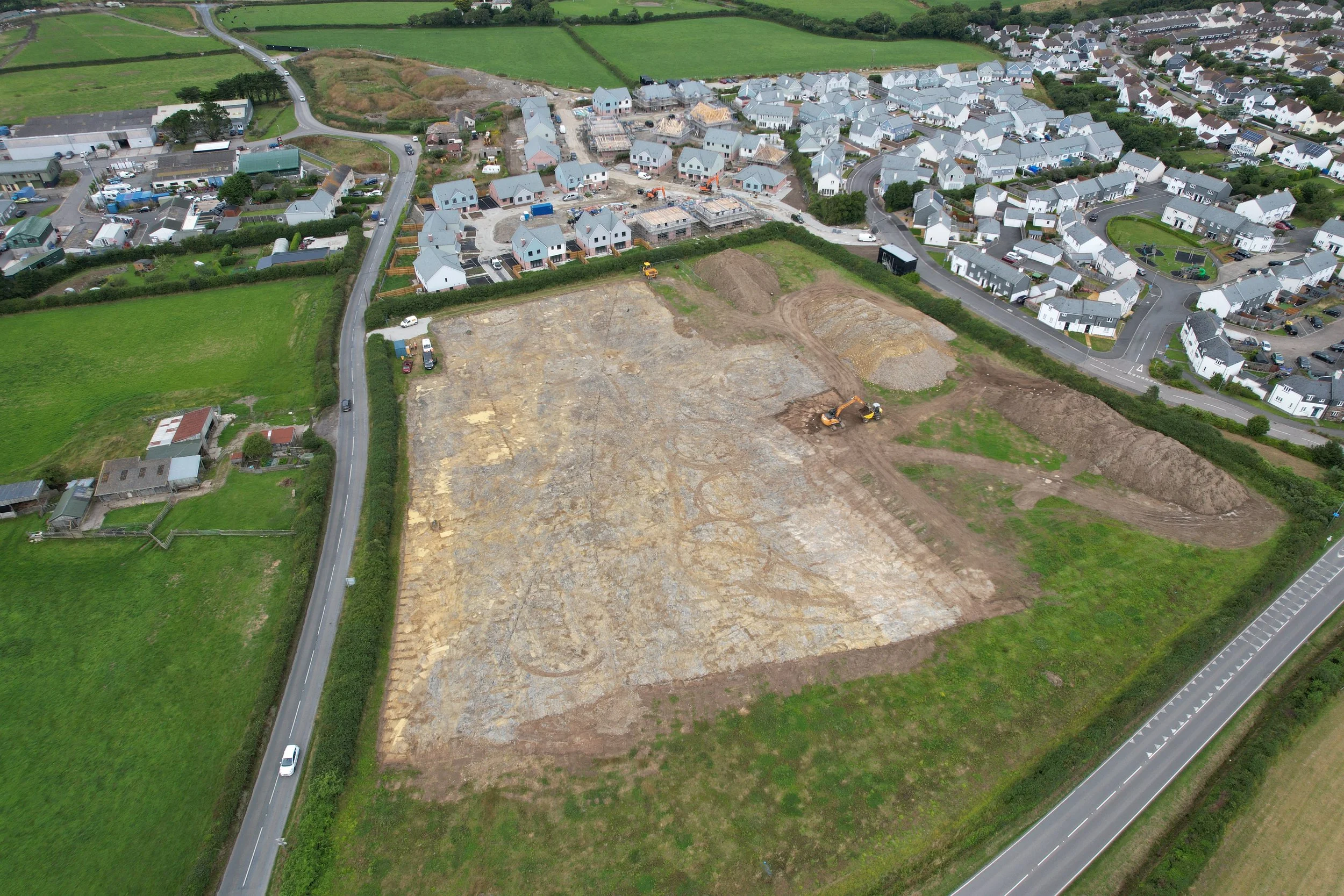 An aerial view of a construction site with excavators working on the soil, surrounded by residential houses and green fields.