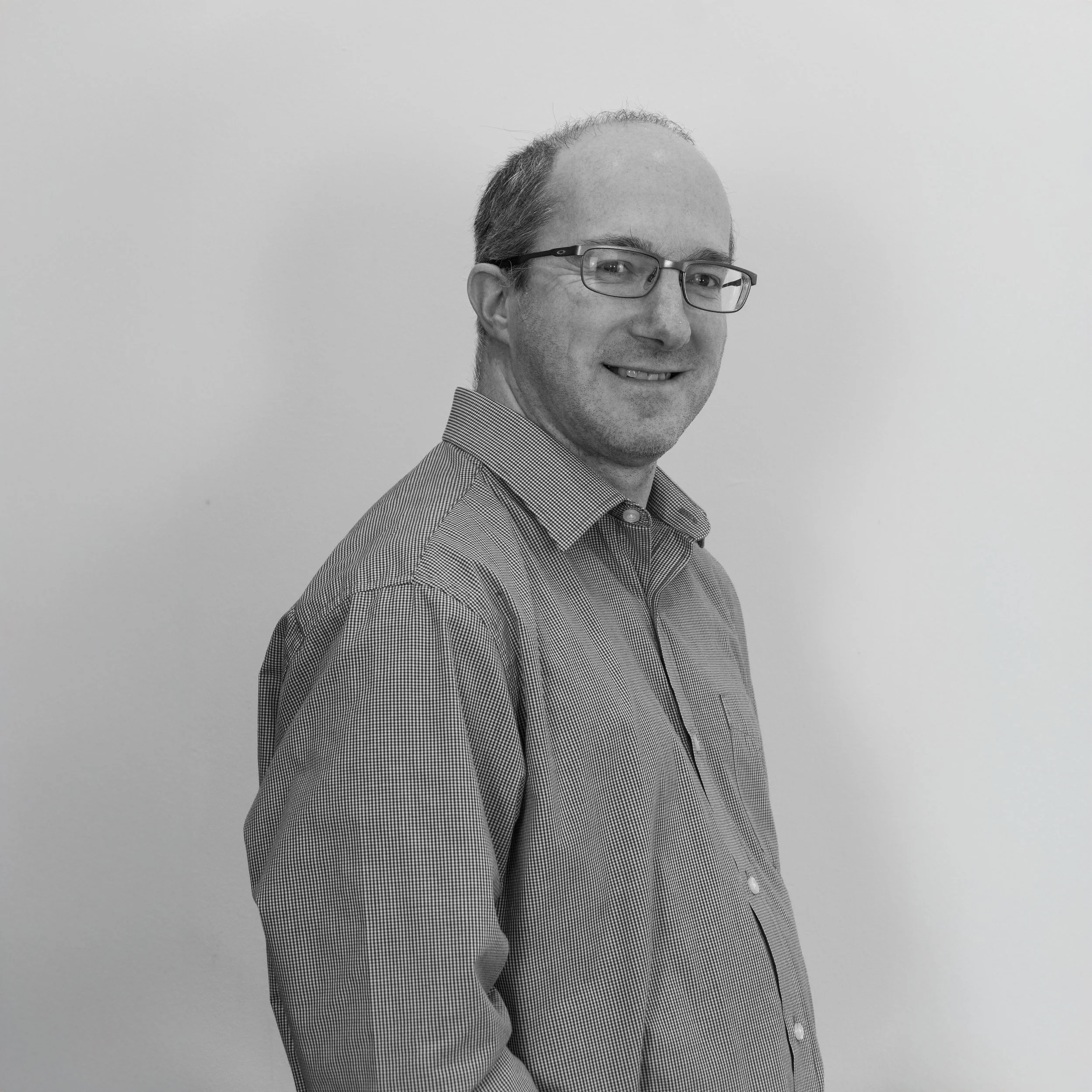 A black and white photo of a smiling man with glasses, wearing a button-up shirt, standing against a plain wall.