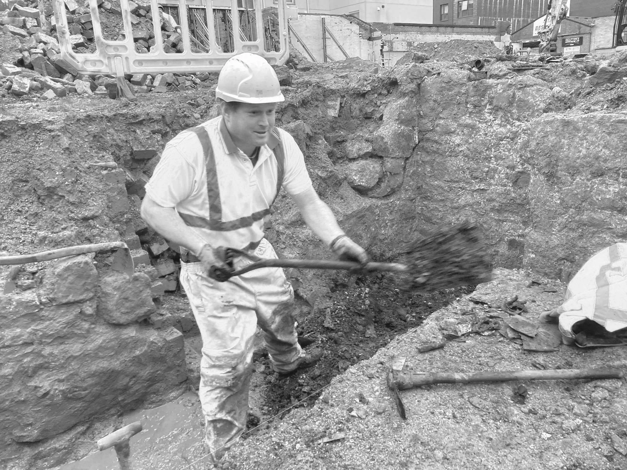 A person wearing a helmet and safety gear is working inside a narrow, rocky underground tunnel, possibly an excavation site, with a bucket nearby.