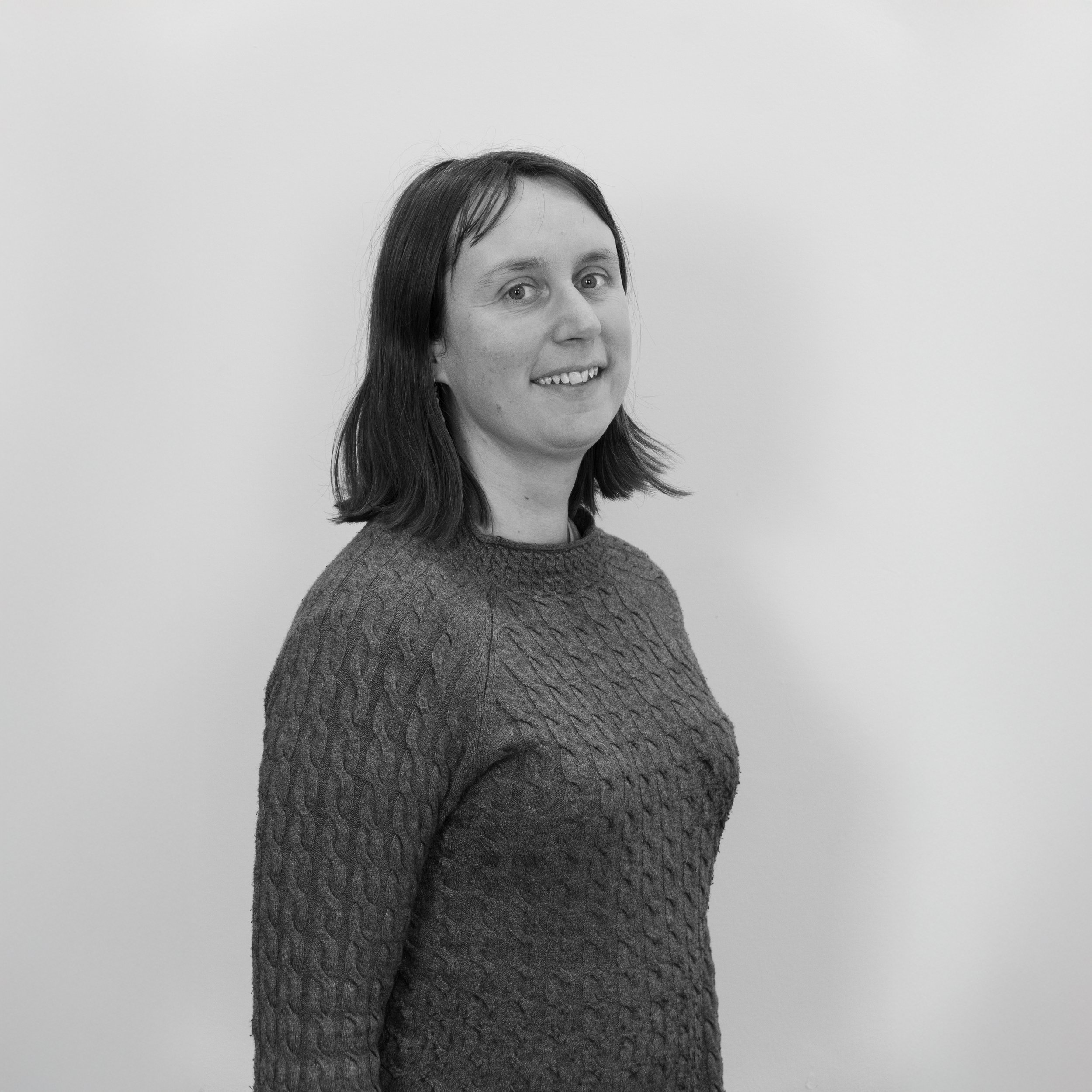 Black and white photo of a woman with shoulder-length dark hair, smiling, wearing a textured sweater, standing against a plain light-colored background.