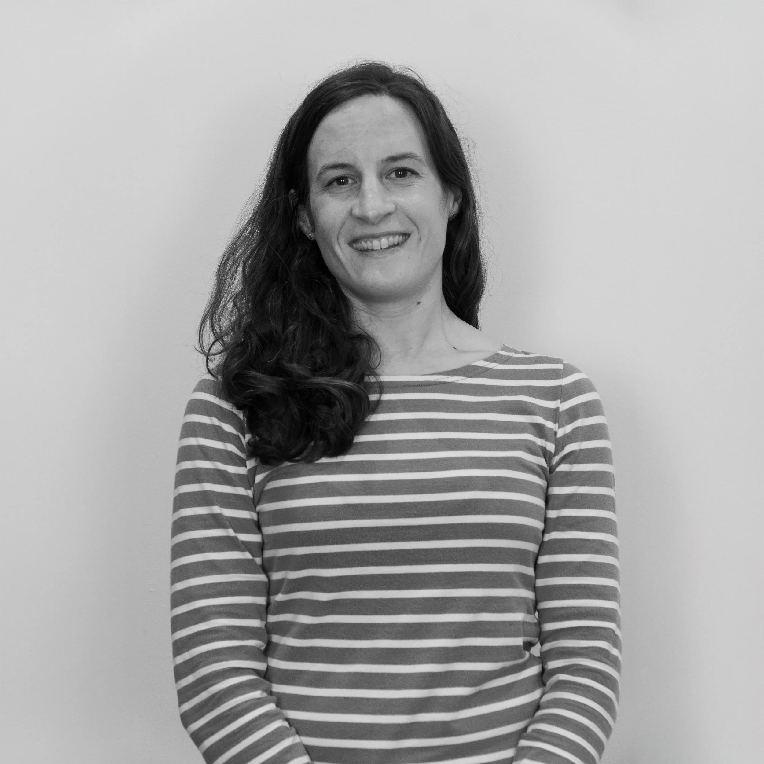 Black and white photo of a woman with long wavy hair, wearing a striped long-sleeve shirt, smiling and standing against a plain background.