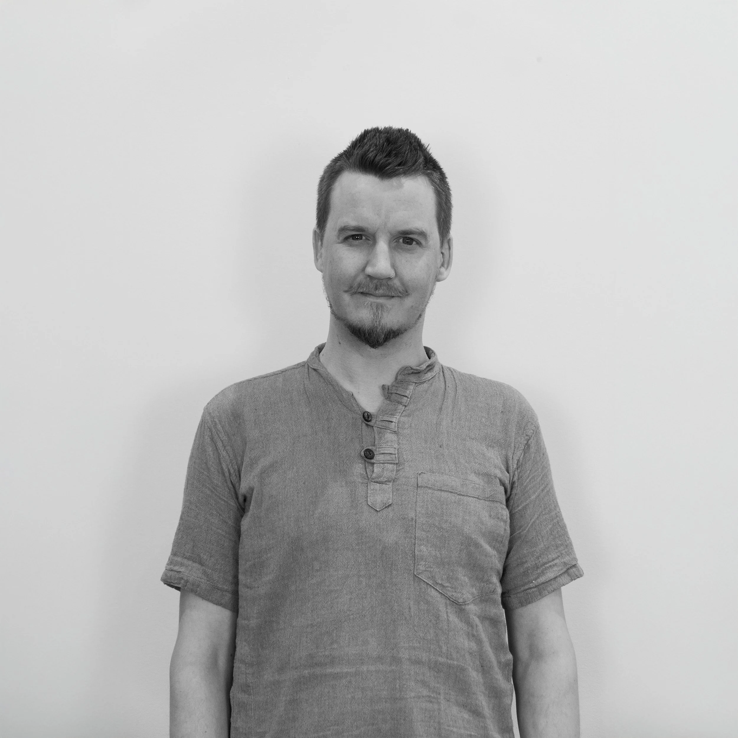 Black and white portrait of a man with short hair and a goatee, wearing a casual short-sleeved shirt, standing against a plain white background.
