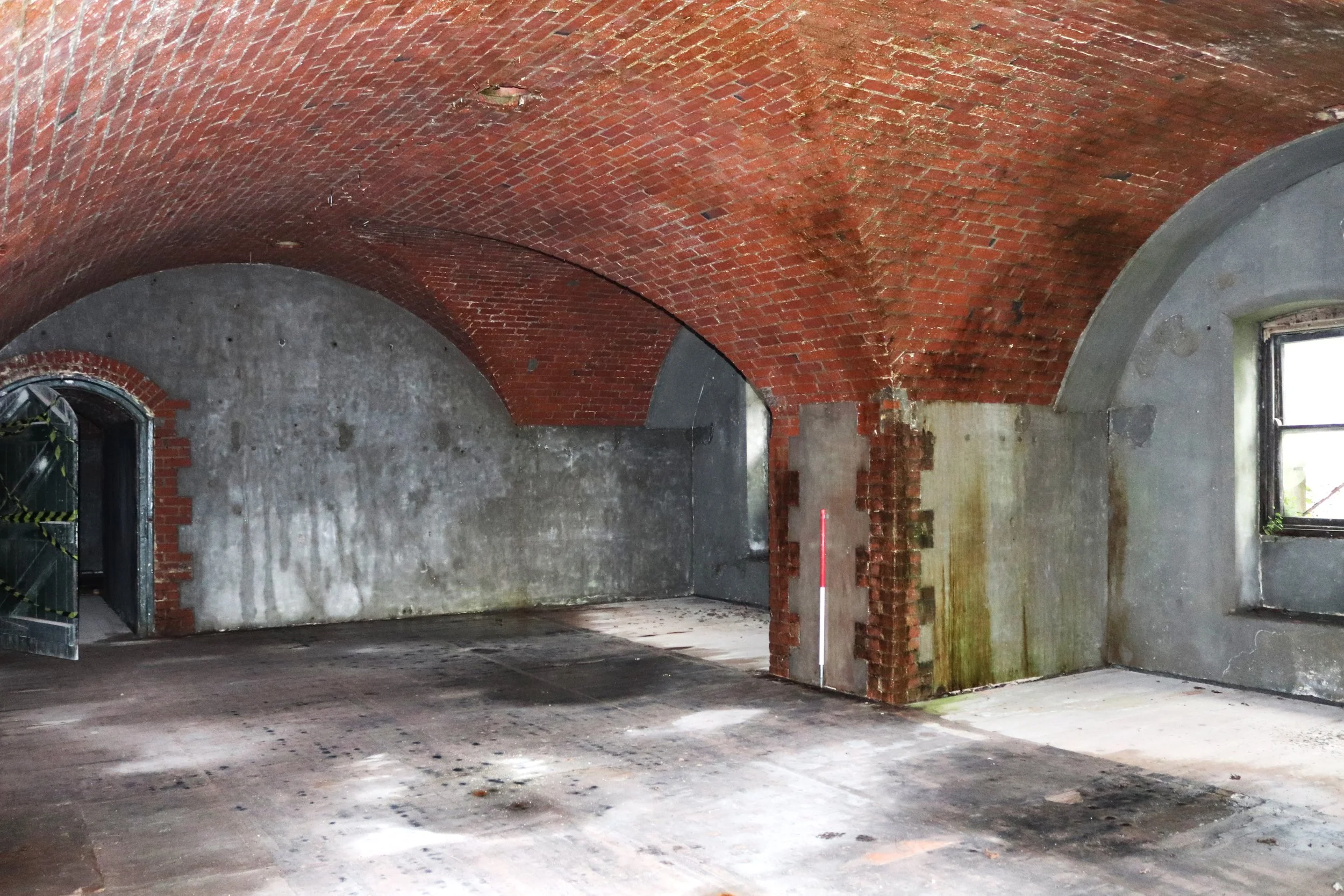An empty, unfinished room with arched red brick ceilings, gray concrete walls, a window with black framing on the right, and a partially constructed brick wall in the center. The floor appears to be concrete with stains.