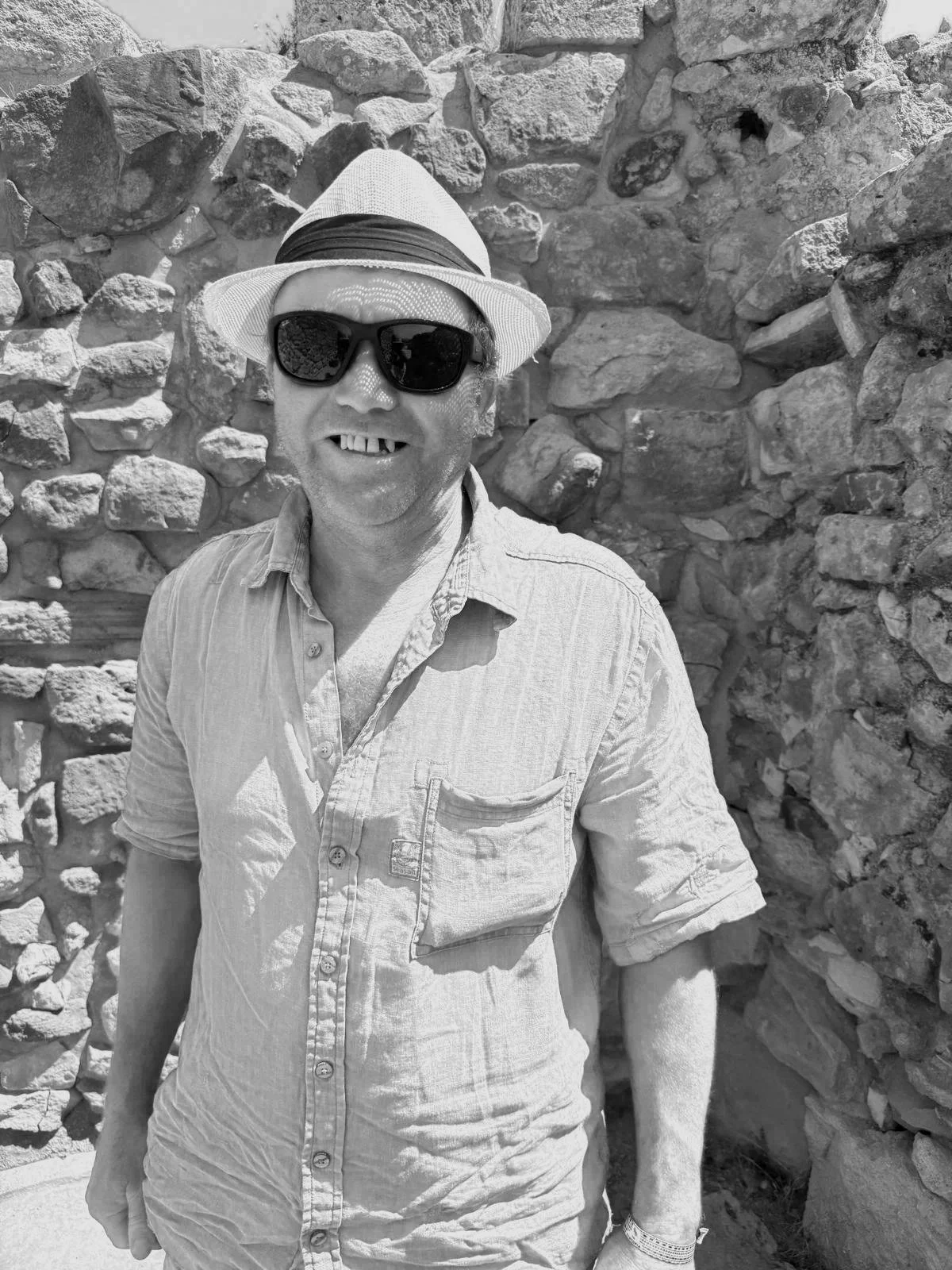 A man wearing a hat, sunglasses, and a short-sleeved shirt is smiling in front of a stone wall.