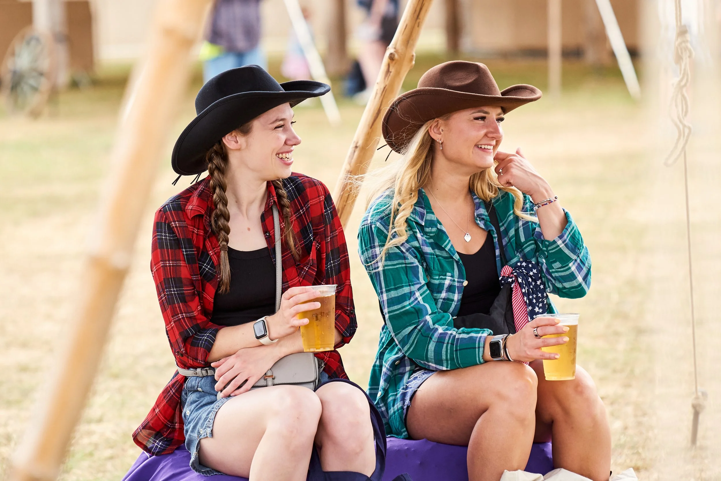 Two young women wearing cowboy hats sitting on a bench at an outdoor event, smiling and holding drinks. One woman wears a red and black plaid shirt, the other wears a green and blue plaid shirt.