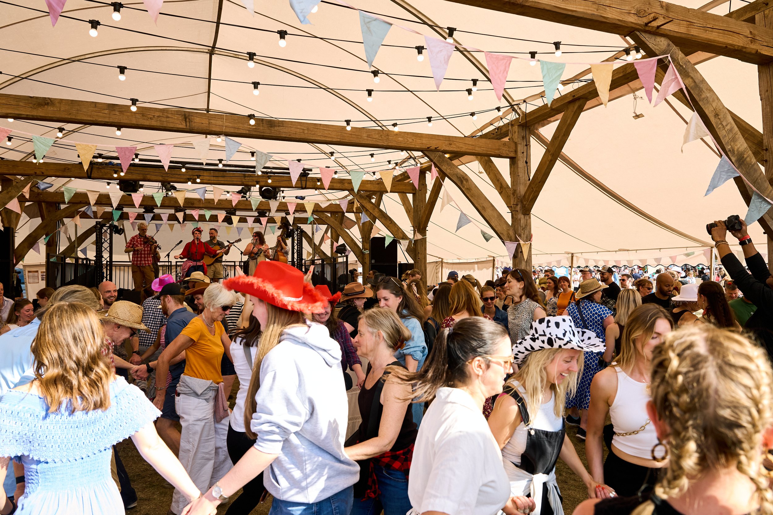 Crowd of people at a festival or outdoor event, dancing and holding hands under decorative string lights and pastel-colored bunting flags, with a band performing on stage in the background inside a large tent with wooden support beams.