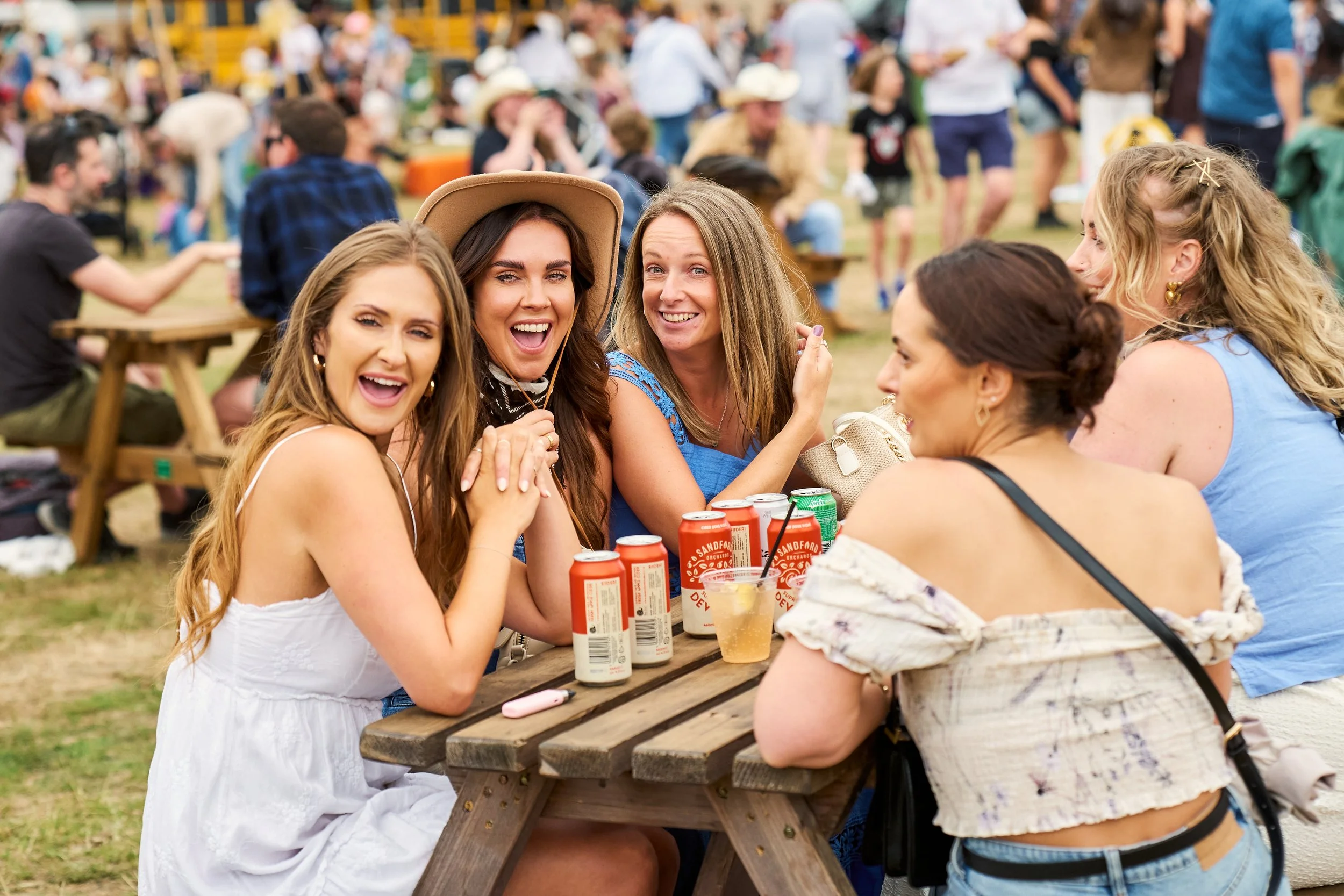 A group of women sitting at a picnic table enjoying drinks and conversation at an outdoor event with many people in the background.
