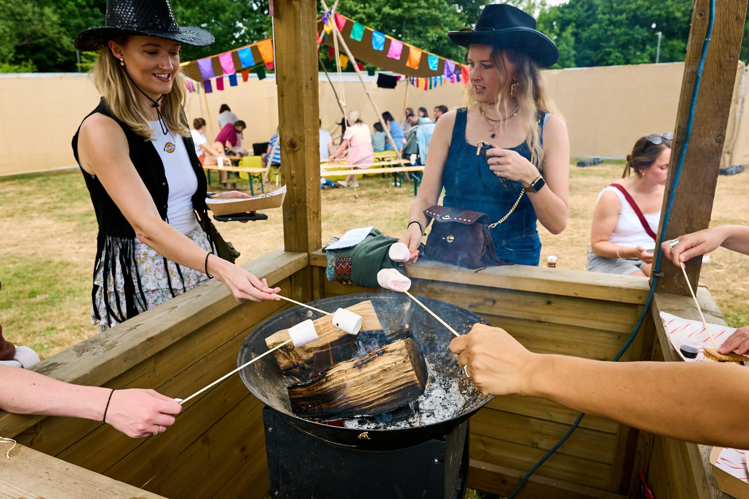 A group of women wearing black cowboy hats gathered around a fire pit, roasting marshmallows on skewers at an outdoor gathering with colorful banners hanging overhead.