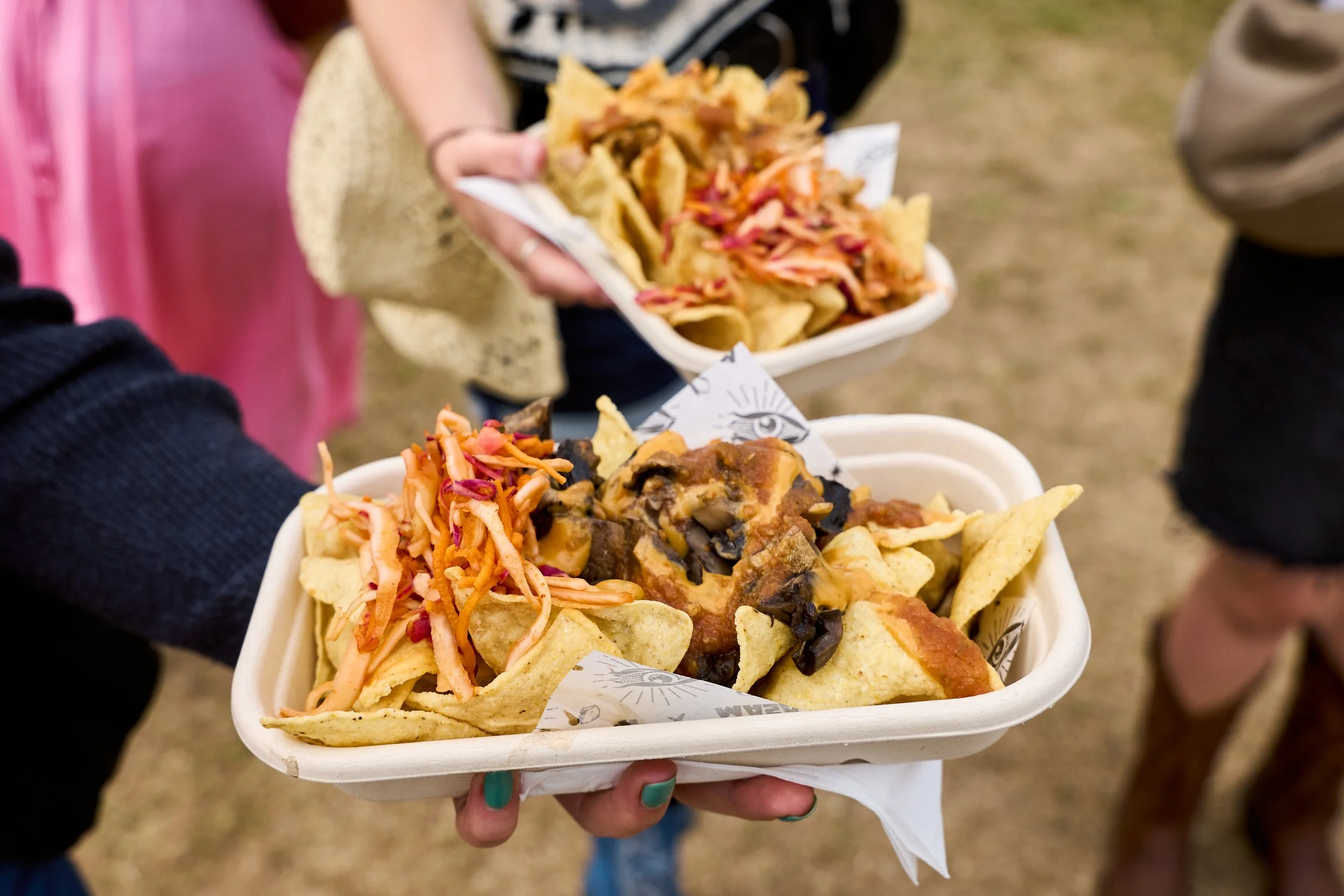 Person holding a food tray with nachos with cheese, pulled pork, salsa, and shredded vegetables, at an outdoor event.