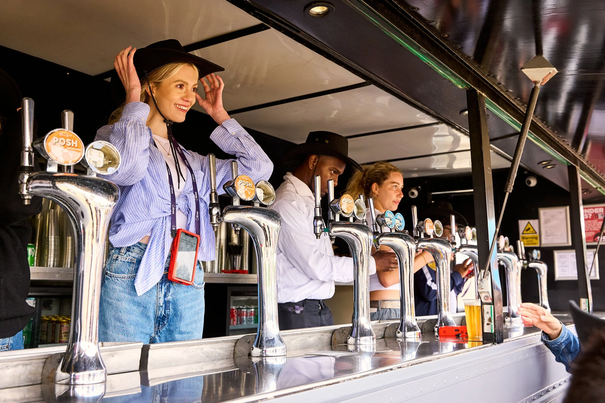 Smiling young woman in striped shirt adjusting her hat while standing at a food or drink stand, with other people behind the counter serving drinks and interacting.