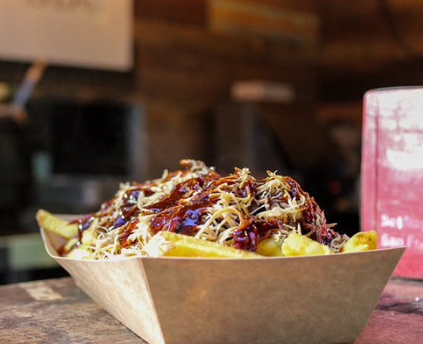 A serving of loaded fries topped with shredded cheese, pulled pork, barbecue sauce, and shredded lettuce in a paper tray on a wooden table.