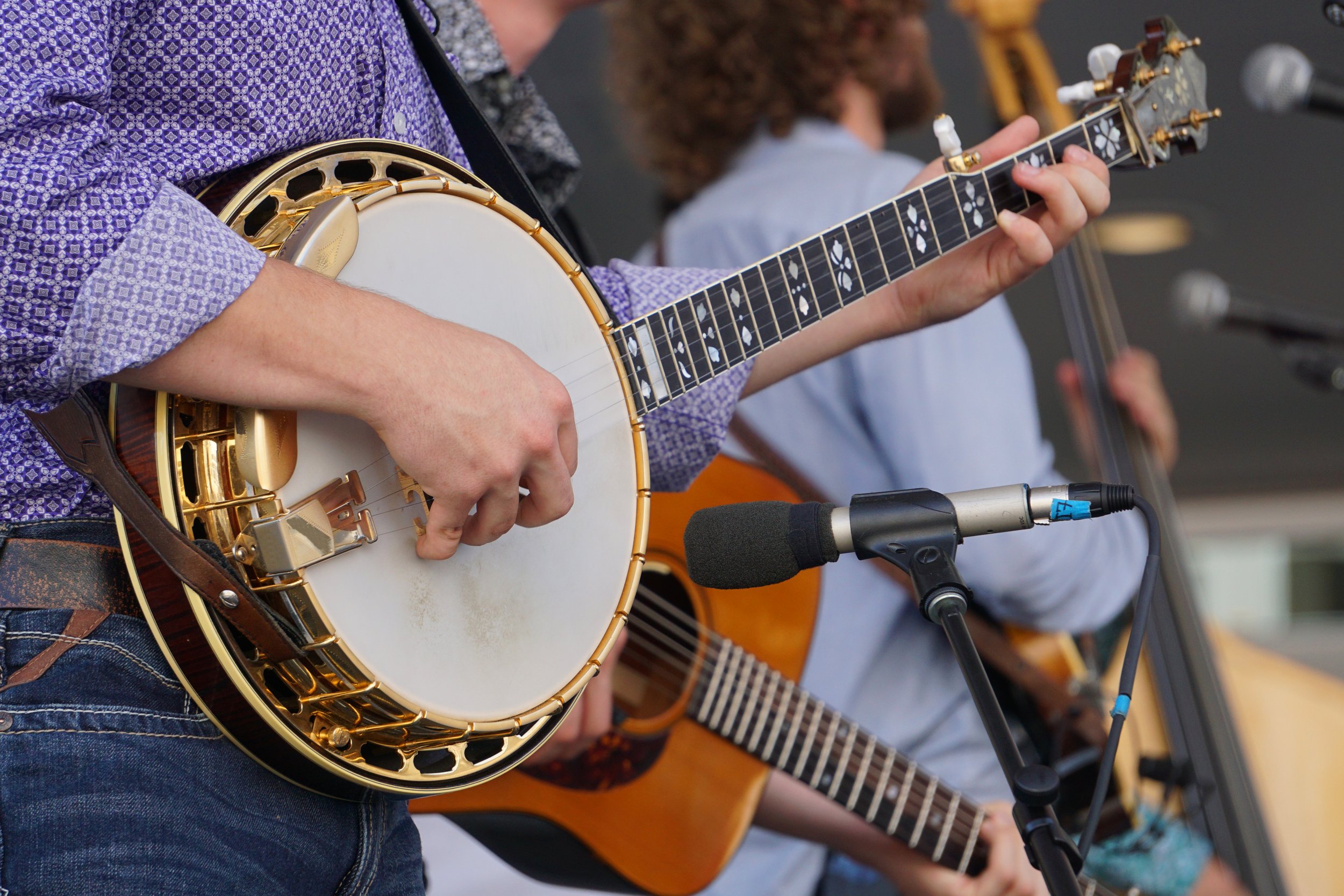 Musician playing a banjo with a microphone; band in background.