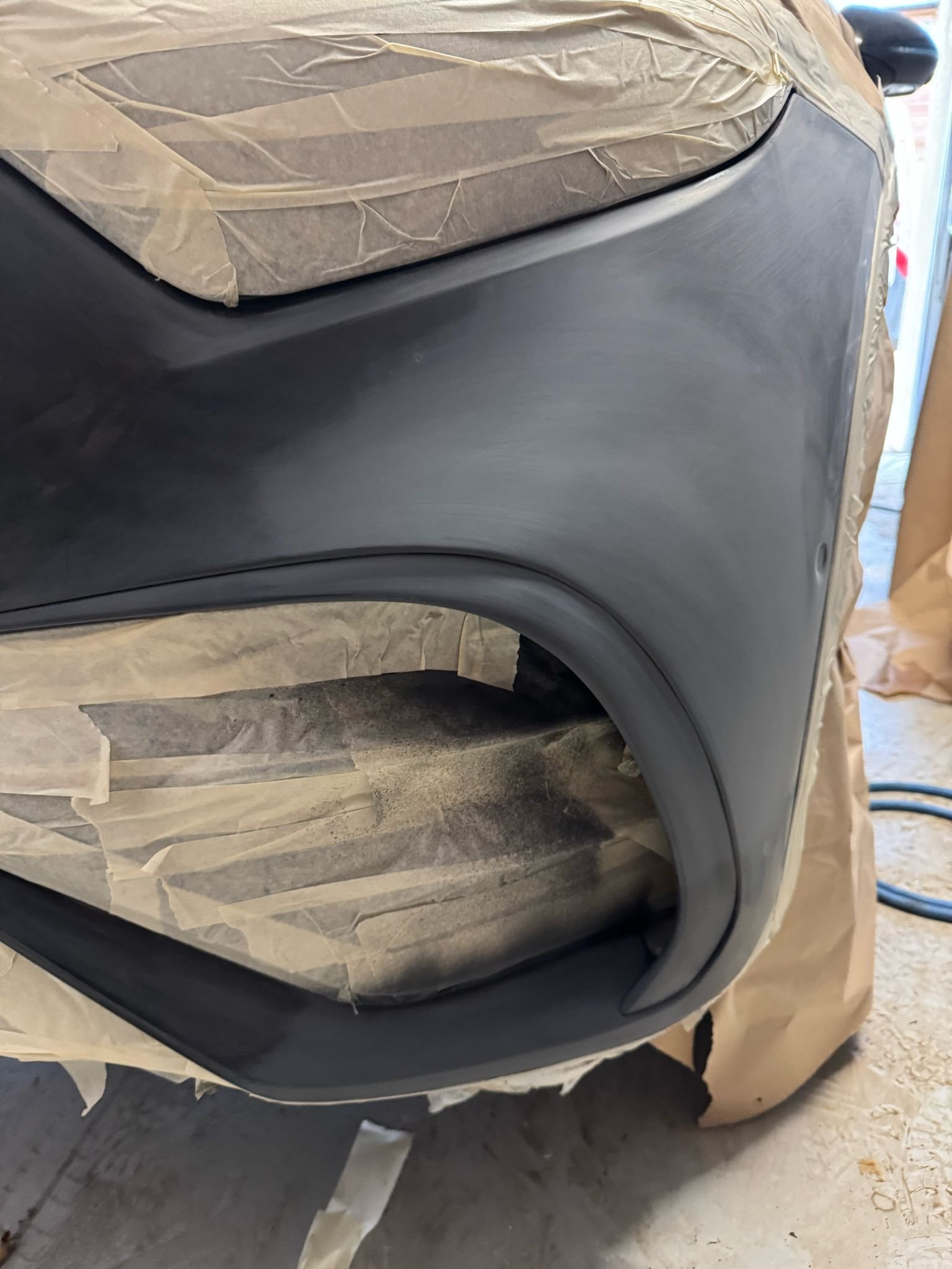 Close-up of a black car bumper being painted, partially masked with masking tape and paper.
