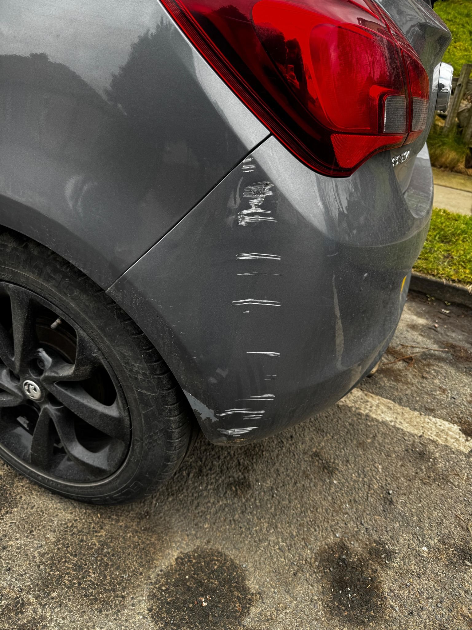 A gray car with visible scratches and scuff marks on the rear left side near the bumper.