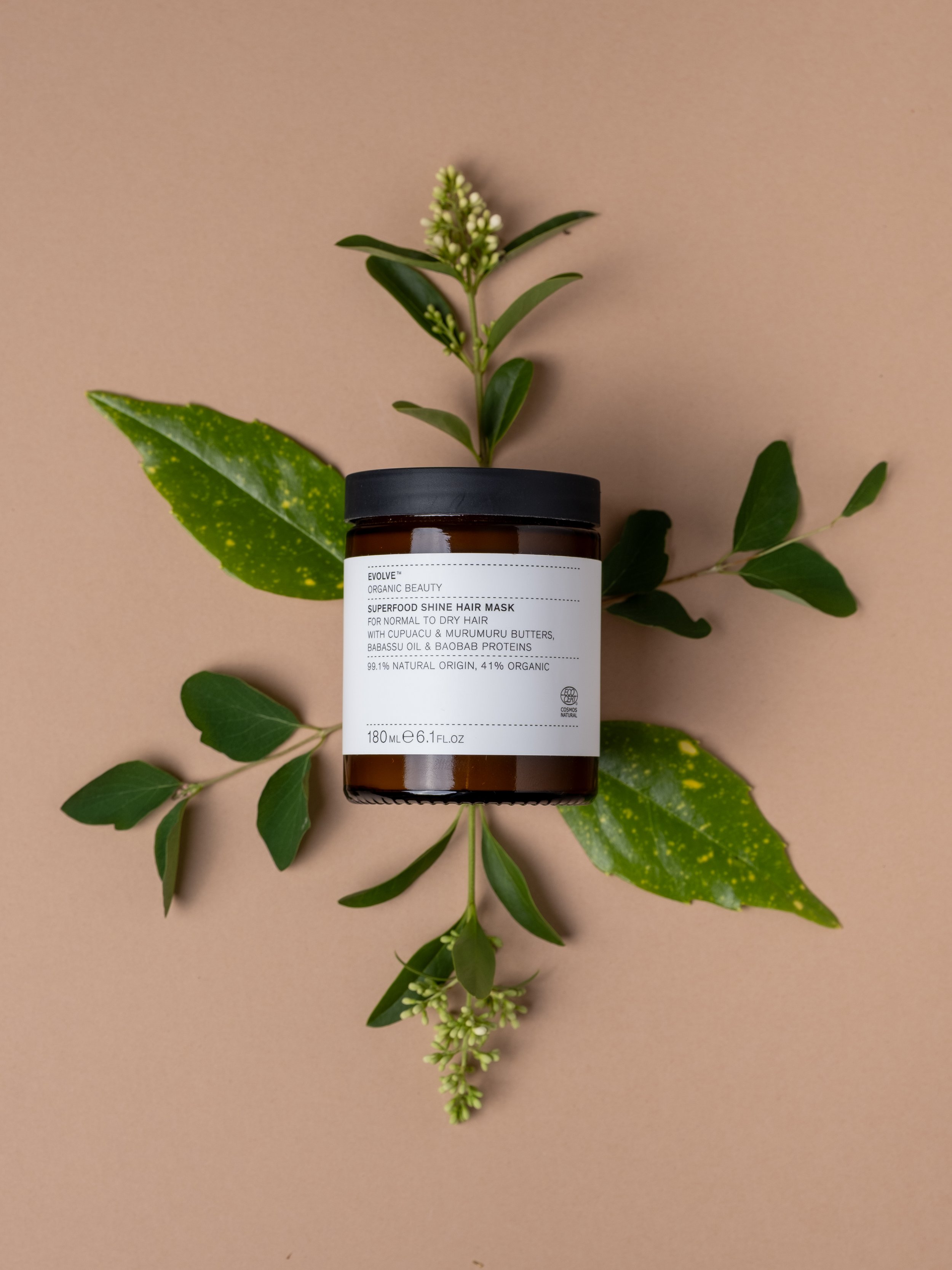 A container of Evolve Superfood Shine Hair Mask placed on a beige background surrounded by green leaves and small white flowers.