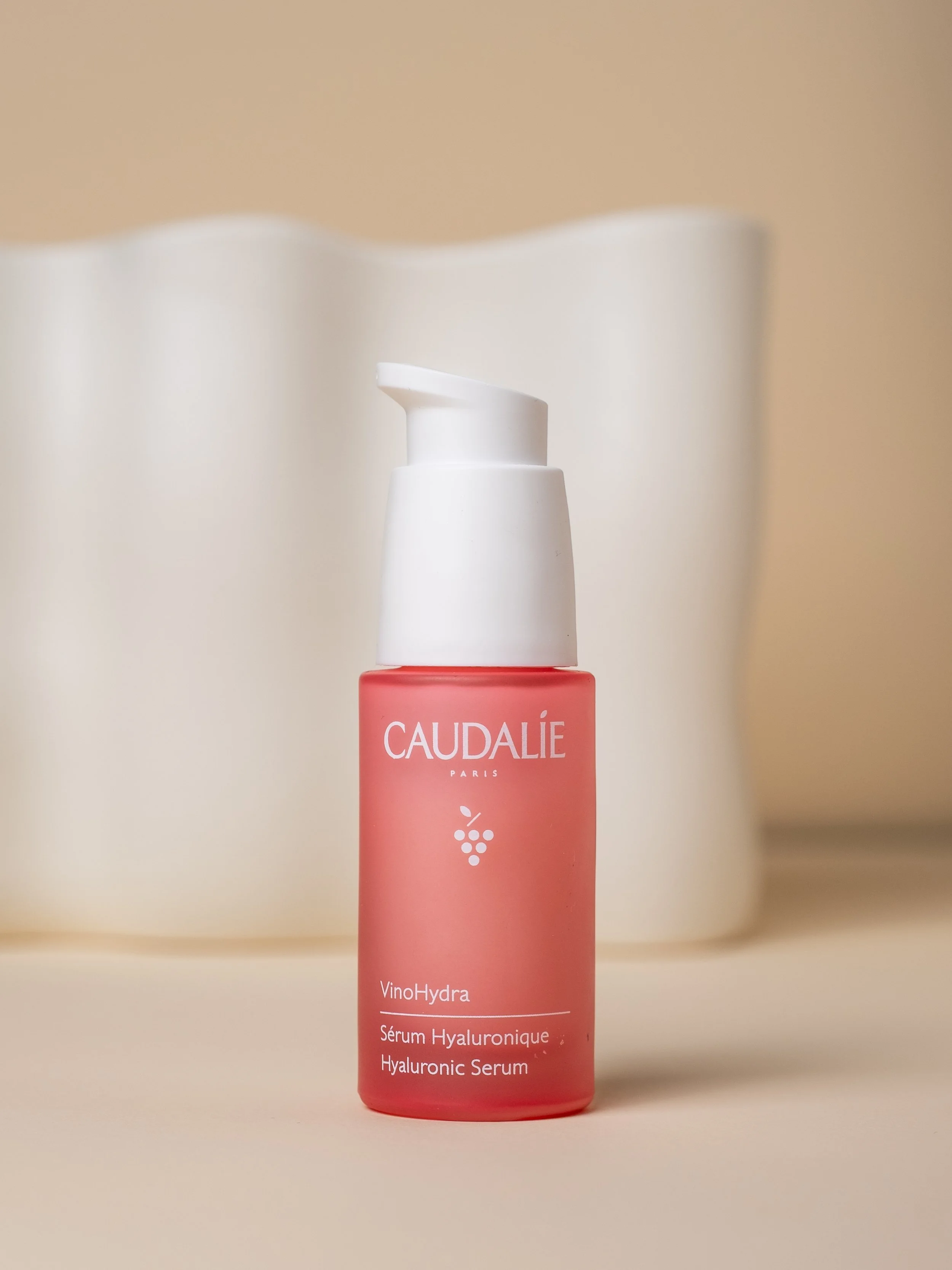 Pink serum bottle on a cream background in front of a white ceramic vase