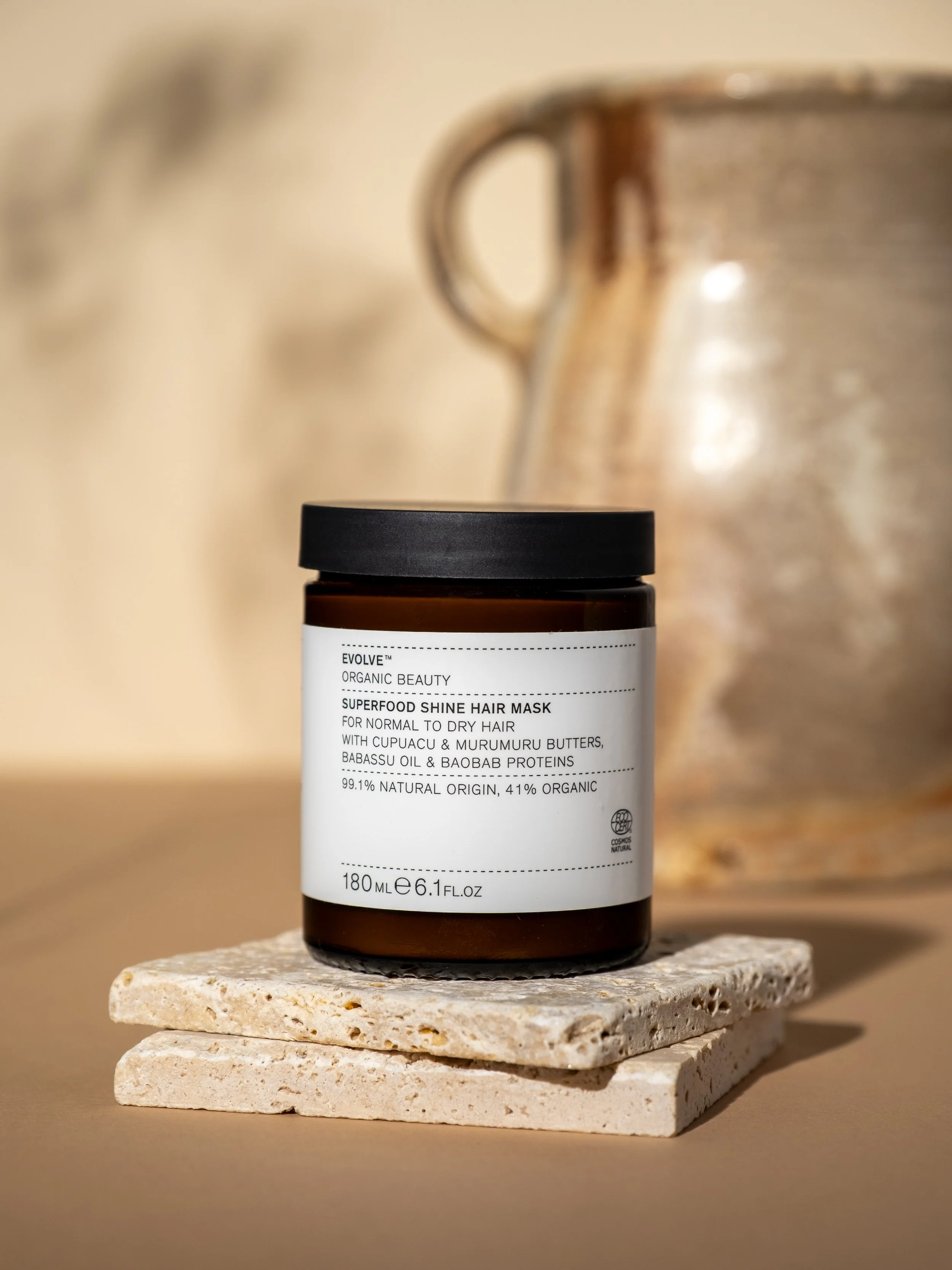 Brown jar with black lid labeled 'Superfood Shine Hair Mask' placed on a stone mini platform, with a ceramic teapot or pitcher in the background.